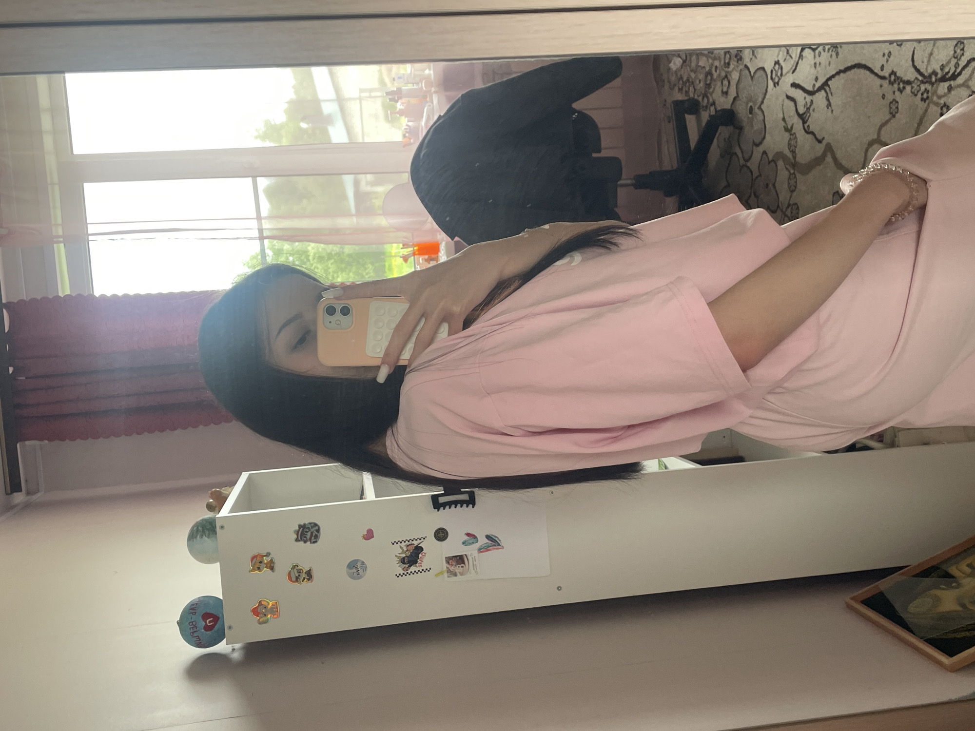 A young woman is captured in a mirror selfie, her reflection showing her from the chest up, angled slightly to the side. She has long, dark hair and is wearing a pink, long-sleeved top. Her right arm is visible, adorned with a light-colored beaded bracelet on her wrist, and her fingernails are painted white. Her face is largely obscured by the light peach-colored smartphone she holds up to take the photo, which features two distinct camera lenses and a textured pattern on its back.
The reflection behind her reveals an indoor setting, likely a residential room. To the left, a large window is visible, letting in abundant natural light and showing green foliage from trees outside, suggesting a bright daytime. A black office-style chair is reflected further to the right. Below her torso, a white, multi-compartment storage unit or shelf is visible, decorated with several stickers. These include characters from "Paw Patrol," a heart, a checkered flag pattern, abstract shapes, and a circular sticker with partially legible text that appears to say "EYE ❤ BELIEF." Two small, spherical objects rest on top of this unit. In the upper right corner of the reflection, a patterned rug or carpet with floral designs in beige and brown hues is visible. The floor in the foreground appears light-colored, and a small framed picture rests on it near the white storage unit.