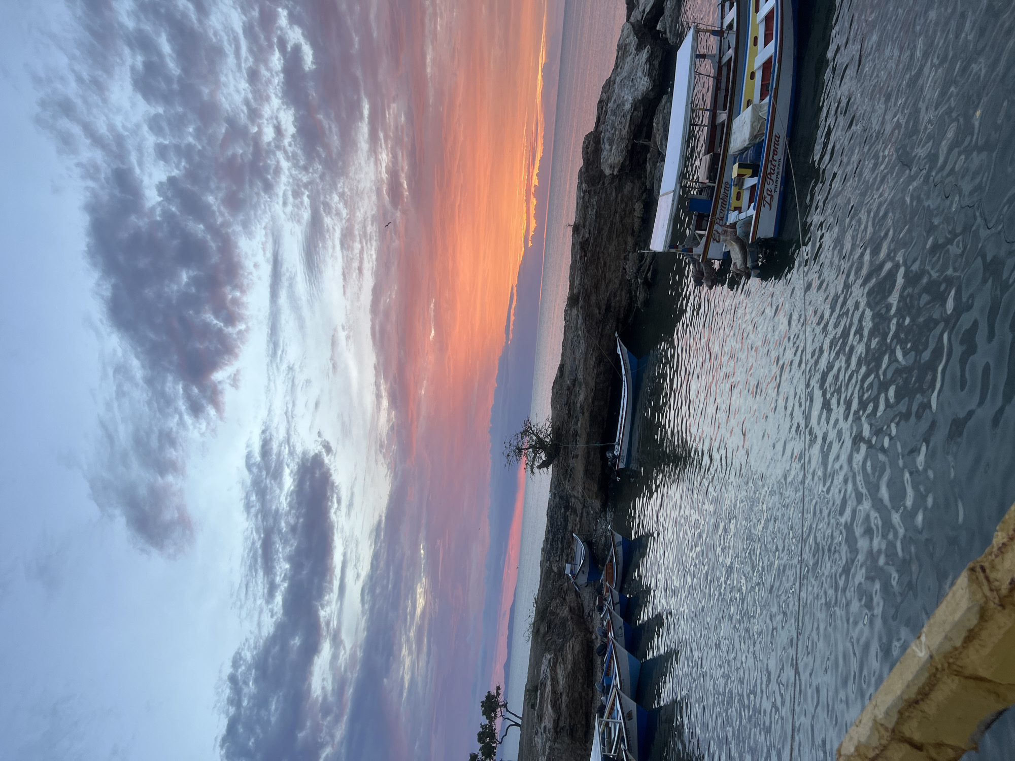 The image depicts a coastal scene in Juan Griego, Venezuela, at dusk. The sky is filled with dramatic clouds, colored with hues of orange, pink, and purple from the setting sun, transitioning to a soft blue at the zenith. The water is calm, reflecting the sky's colors and the surrounding environment.
Several small boats are moored in a sheltered cove. These boats are diverse in color and design, with some featuring white hulls and colored trim, while others are primarily white with red and yellow accents. One boat is labeled "La Patrona." Another appears to be named "El Bambino." The boats are tied with ropes to the rocky shore.
The shoreline is characterized by a rugged, rocky formation interspersed with sparse vegetation. Some trees with dark foliage grow on the rocks. In the distance, a mountain range is visible against the horizon, with the sea stretching out to meet the sky. A yellow-topped structure, possibly a railing or a portion of a dock, is visible in the foreground on the lower left. The overall atmosphere is serene and picturesque, characteristic of a tropical sunset.