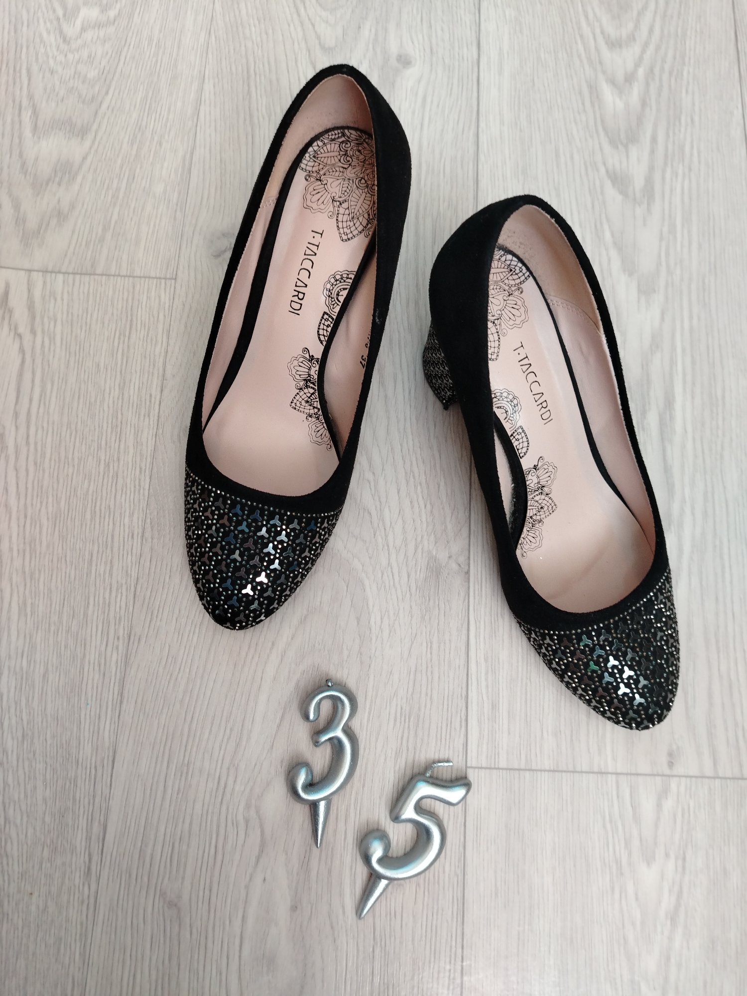 A top-down shot captures two black women's shoes and two silver number candles laid out on a light-colored wooden floor. The setting appears to be indoors in Chaikovsky, Russia.
The shoes are black, possibly made of velvet or suede-like material, with a low heel or flat design. The toe area of both shoes is intricately embellished with numerous small, shiny, silver and iridescent sequins or glitter. The interior lining of the shoes is a light pinkish-beige color, featuring elegant black lace-like patterns or mandalas. Visible text on the insole of both shoes reads "T. TACCARDI". Additionally, the number "37" is partially visible on the insole of the shoe on the right, likely indicating the shoe size.
Below the shoes, two silver numeric candles are placed. One is shaped like the number '3' and the other like the number '5'. Both candles have pointed bases, suggesting they are designed to be inserted into a cake or similar item. The overall arrangement of the items implies a display or preparation related to a celebration, possibly a birthday, with the numbers '3' and '5' potentially signifying an age (e.g., 35) or a date. The wooden floor planks have a visible grain and a light, neutral tone.