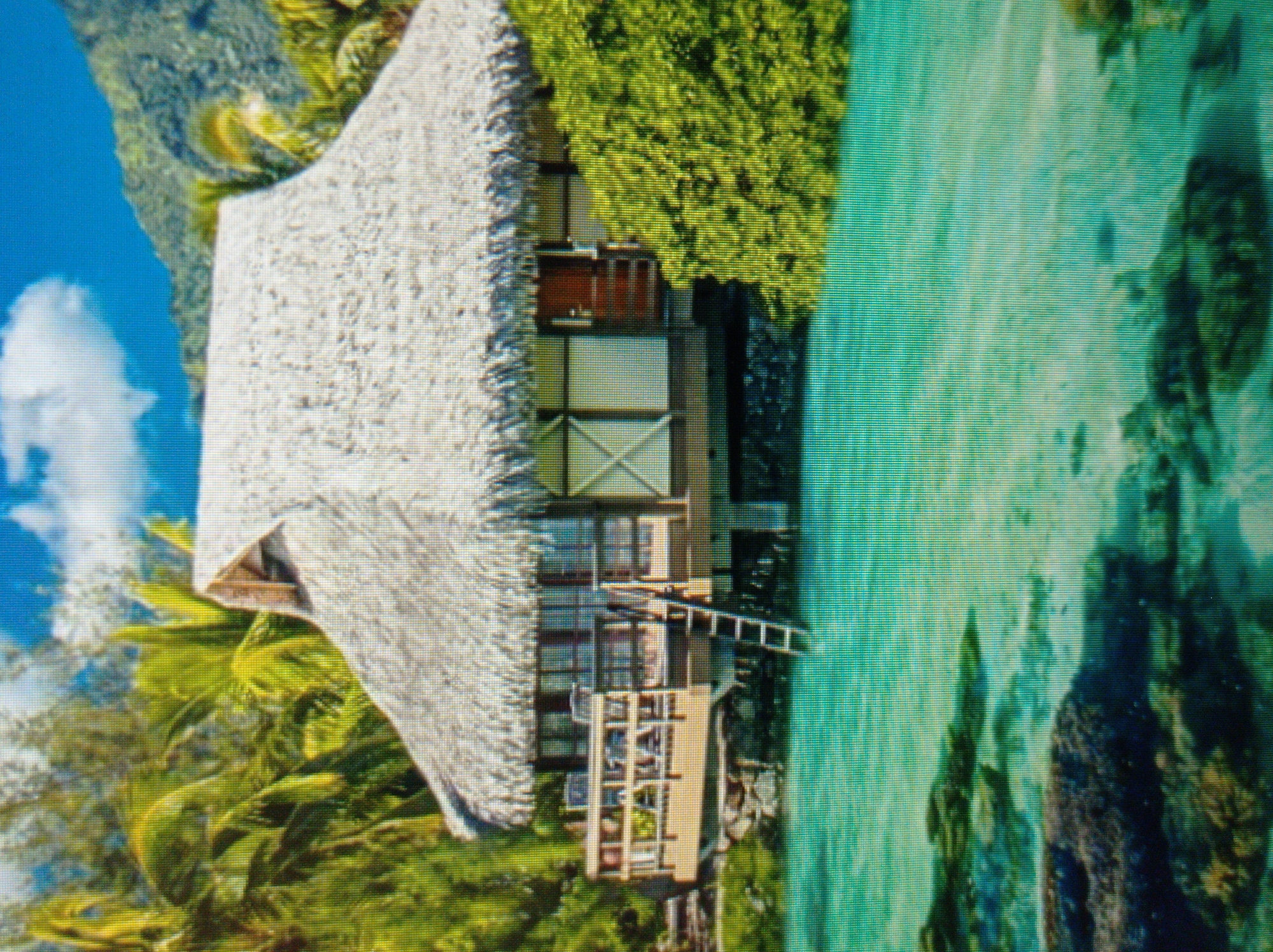 The image depicts an overwater bungalow with a thatched roof, situated in clear turquoise water. Palm trees and lush green foliage surround the bungalow, with a mountainous backdrop under a bright blue sky with scattered clouds. A wooden deck with railings extends from the bungalow, featuring a ladder leading down to the water. The water is shallow enough to reveal rocks and coral formations beneath the surface. The scene suggests a tropical or subtropical resort location. There are no people present in the image. The image provided does not contain any information or visual cues to suggest a location in the city of Mukachevo, Ukraine.