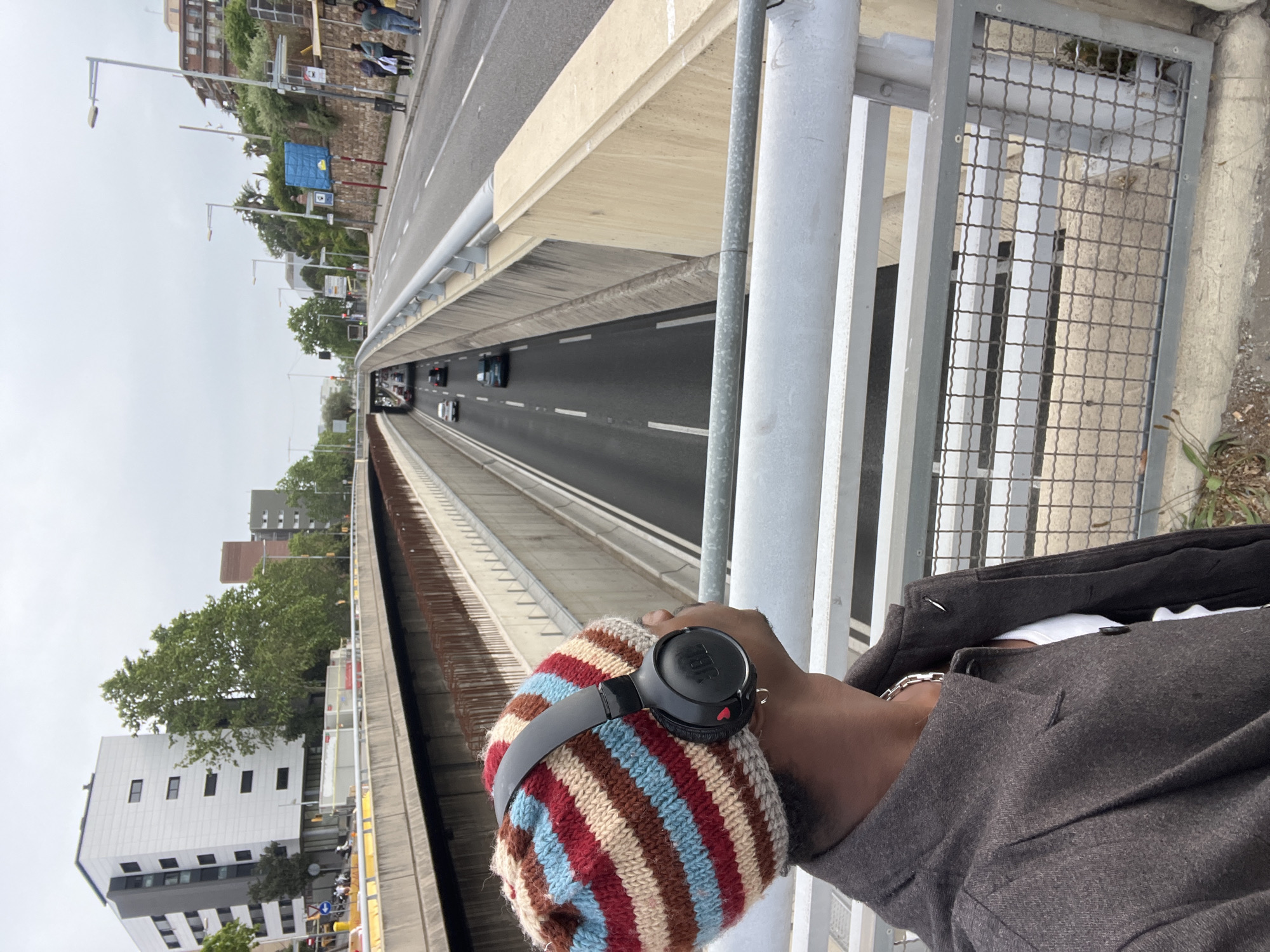 The image, rotated 90 degrees clockwise, presents an overhead view of an urban landscape in Barcelona, Spain, captured from an elevated perspective such as a bridge or overpass. In the foreground, partially visible on the right, is a person wearing a multi-colored striped beanie with red, blue, brown, and beige hues, and black JBL over-ear headphones. A dark, possibly tweed or wool jacket covers their torso, revealing a silver chain around their neck. The person's hand appears to be gripping a grey metal railing that runs along the overpass.
Below the overpass, a multi-lane highway or major road stretches into the distance, with several cars traveling in both directions. The sky overhead is overcast, indicating a cloudy daytime setting. To the left of the highway, an urban environment features various buildings, including older brick structures and a modern white building with dark-framed windows. Green trees are interspersed among the buildings and along the roadside. Streetlights line what appears to be an upper road or pathway parallel to the highway, where a few people can be seen walking in the distance. Construction barriers and other street furniture are also visible in this area. The only discernible text is the "JBL" logo on the headphones.