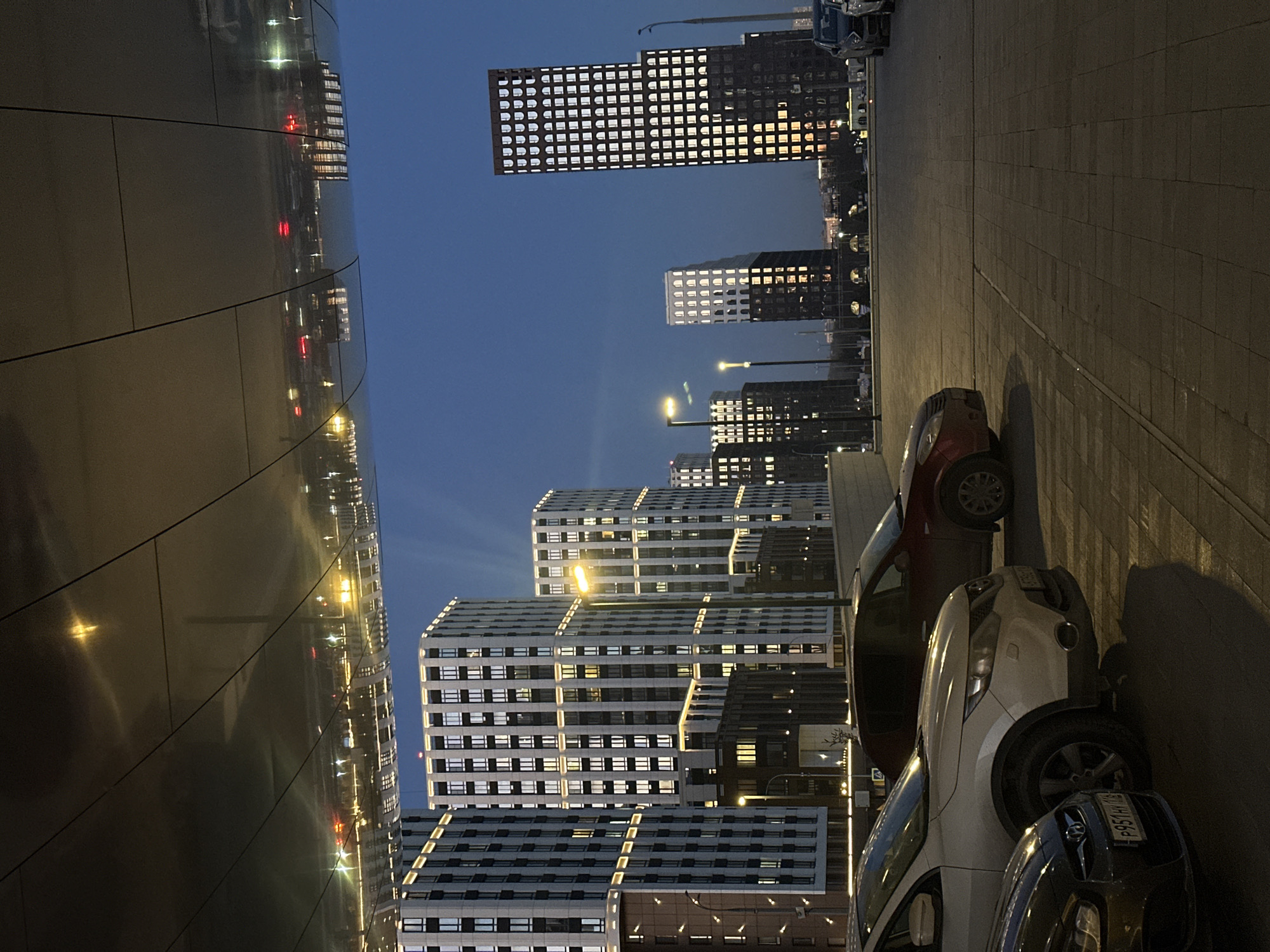 The image presents an urban night scene in Kazan, Russia, characterized by modern architecture and parked vehicles under a dark, clear sky. The time of day is dusk or night, evident from the illuminated buildings and streetlights.
On the left, a prominent, curved building with a highly reflective, metallic facade runs vertically, reflecting the bright lights of the opposite structures and streetlights in distorted patterns across its surface.
Across the street, a complex of tall, contemporary buildings dominates the background. Some are dark-colored, likely black or dark grey, featuring a grid of numerous square windows, many of which are brightly lit. Other buildings are lighter, appearing white or light grey, and exhibit a distinct stepped or staggered design with many lit windows and glowing architectural lines along their ledges. The overall architectural style is geometric and modern.
In the foreground on the right, several cars are parked on a paved area, suggesting a street or plaza. These include a dark red car, a white SUV or crossover, and the front portion of a dark blue car. The license plate of the dark blue car is partially visible, displaying "P951HY" and the country code "RUS", affirming the Russian location context.
No people or specific activities are visible, depicting a static urban environment after dark. The illuminated windows suggest habitation or ongoing activity within the buildings, contributing to a sense of a bustling city at rest.