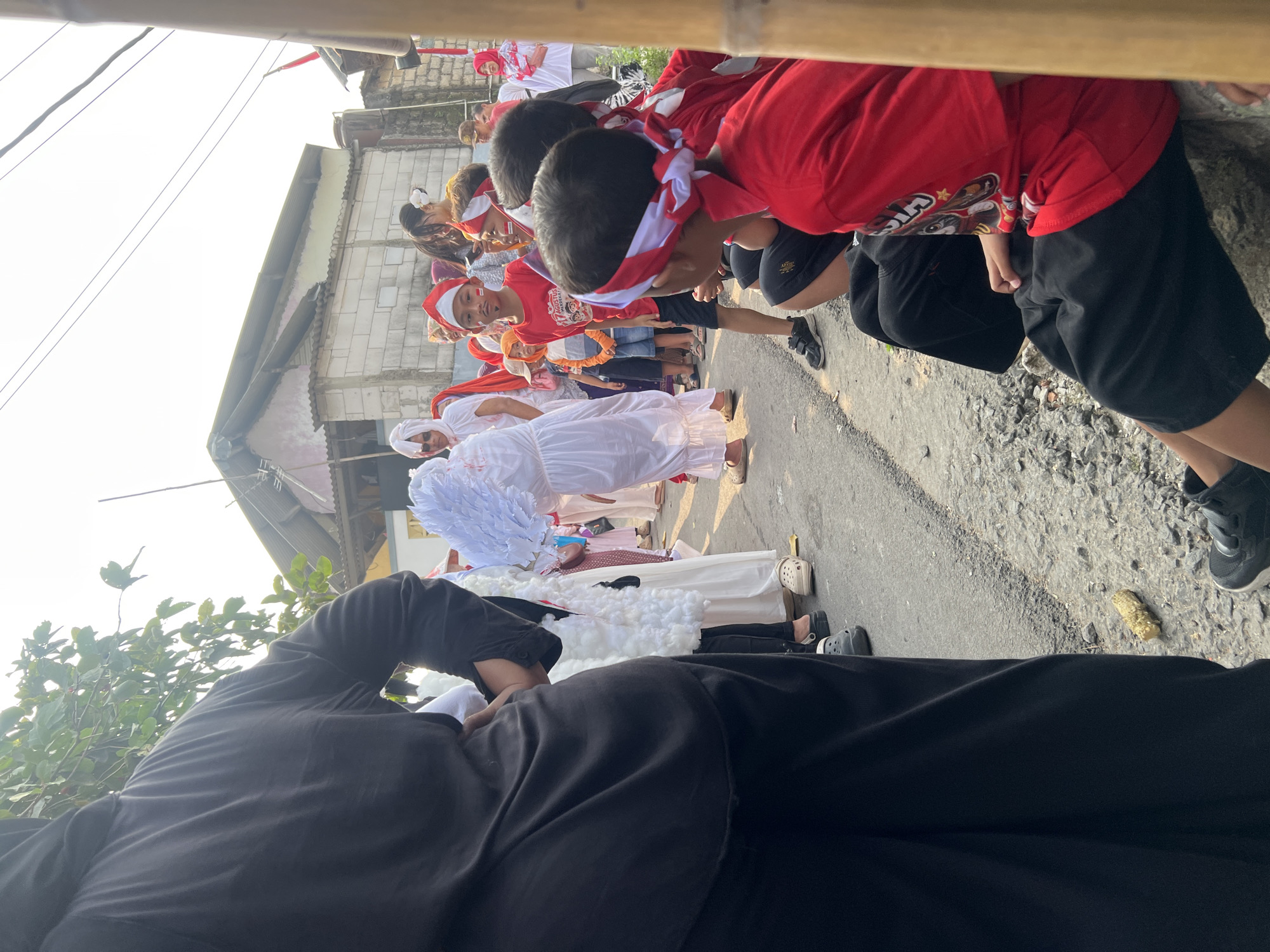 This image captures a street scene in Klapanunggal, Indonesia, likely during a celebratory event. In the foreground, a person wearing a black abaya stands with their back to the camera, obscuring much of the left side of the frame. To their right, a line of children and adults are visible, many dressed in red and white attire, suggesting a patriotic theme. Several children wear red shirts and headbands, some with white or red and white stripes, and some have red and white face paint. One young boy in a red shirt and a Santa-style hat stands out. Behind them, a building with a light-colored brick wall and a corrugated metal roof is partially visible. The ground is a paved road with a rough, stony edge. The lighting suggests it is daytime, with bright sunlight. A bamboo pole is visible on the far right of the frame.