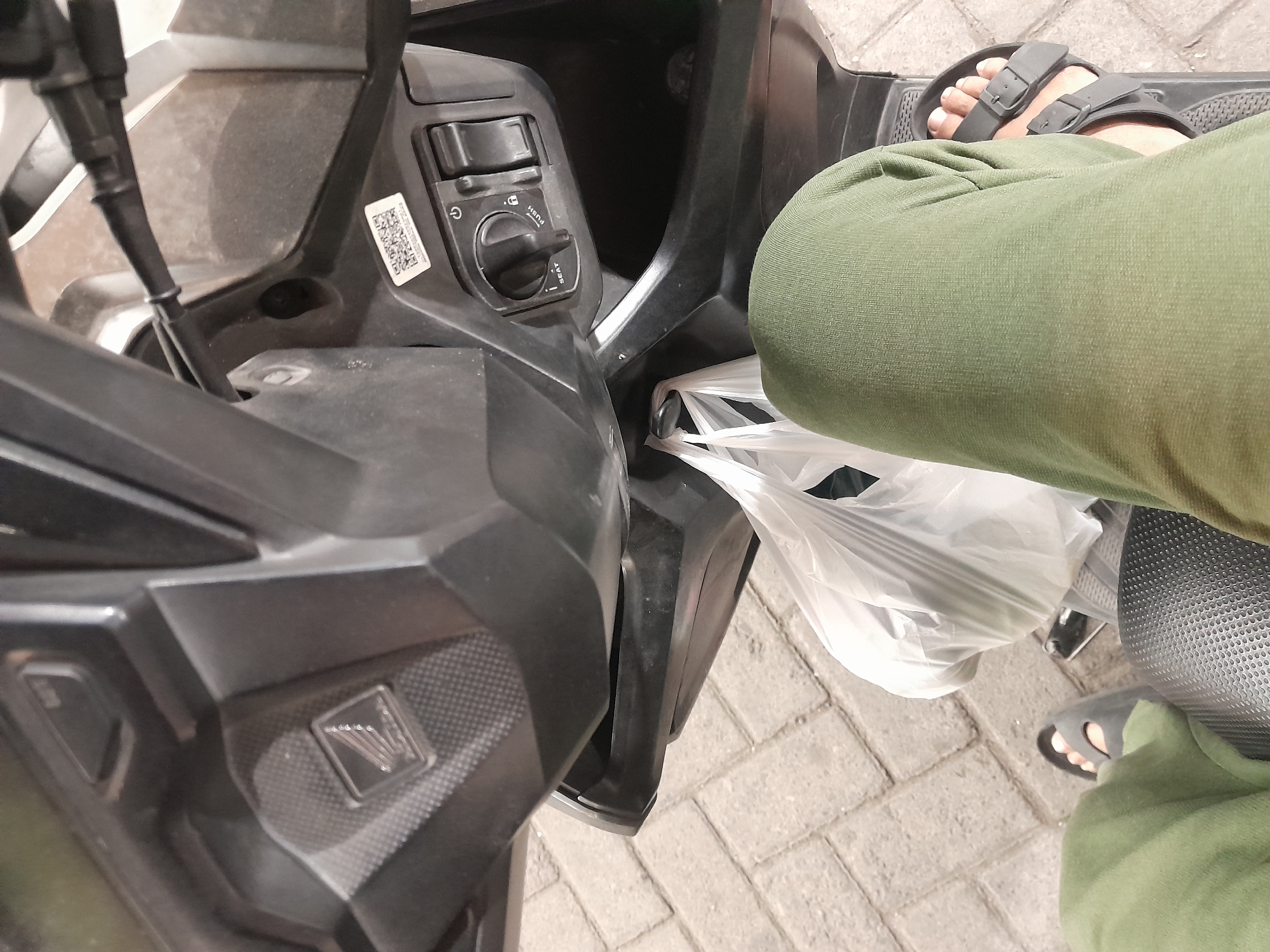 In Desa Ponokawan, Indonesia, a first-person perspective shows a person seated on a motorcycle. The person wears green pants and dark sandals. A white plastic bag containing items is visible hanging from the front of the motorcycle, between the rider's legs. The motorcycle's dashboard is in view, featuring a key ignition with markings for "SEAT" and "PUSH," as well as a power button and a lock symbol. A QR code sticker is also present on the dashboard. The background consists of paved ground, appearing to be a walkway or road made of interlocking bricks.