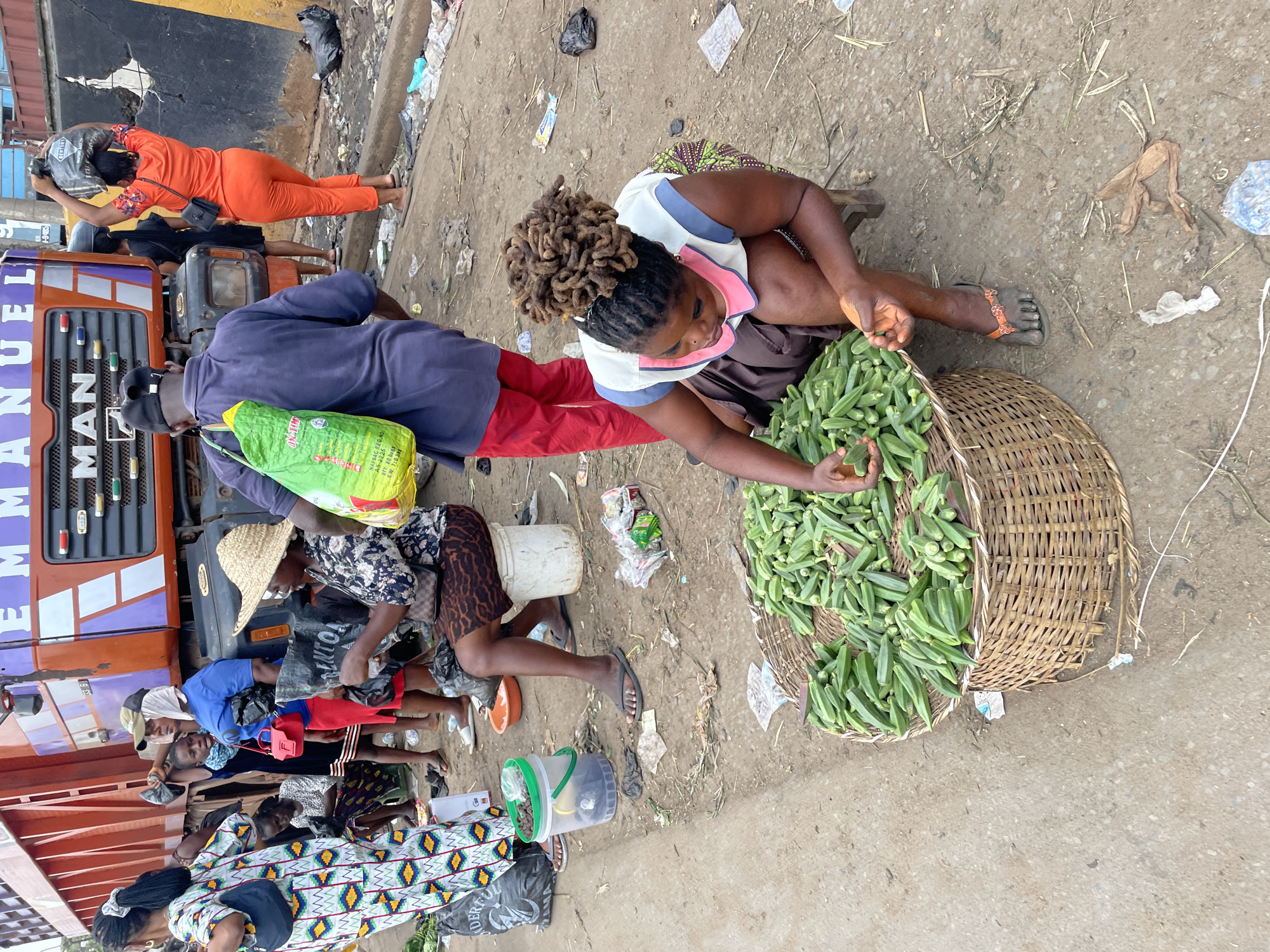 The image depicts an outdoor market scene, likely in a city in Unknown. In the foreground, a woman with dreadlocks is crouched beside a large woven basket overflowing with okra. She is wearing a light-colored top with a pink collar and a dark skirt. Her hands are engaged with the okra. To her left, a clear plastic bucket with a green lid contains what appears to be more produce. Behind her, a man in a dark blue shirt and red pants is walking away from the camera, carrying a green plastic bag over his shoulder. Other people are visible in the background, including women in colorful dresses and headscarves. A large truck with the word "EMMANUEL" on its side is parked behind the people. The ground is dirt and debris-strewn, with various plastic bags and wrappers scattered around. The overall atmosphere suggests a busy, informal marketplace.
