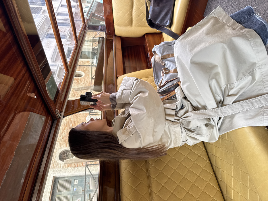 A young female with long, straight, dark brown hair is seated inside a water taxi in Venice, Italy, holding a black compact digital camera to her eye, aiming it out the window. She is positioned in profile, looking towards the right side of the frame. She wears a light beige trench coat tied at the waist, a grey ribbed sweater, and blue jeans. The interior of the boat features glossy dark wooden paneling on the ceiling and upper walls, and quilted mustard-yellow upholstery on the seats. A grey duffel bag with light brown leather straps and trim rests on the seat next to her, and a dark-colored bag with a grey strap is partially visible on an adjacent seat. Through the boat's windows, typical Venetian architecture is visible, including brick buildings with arched and rectangular windows, some with dark shutters, and metal railings, under an overcast sky.