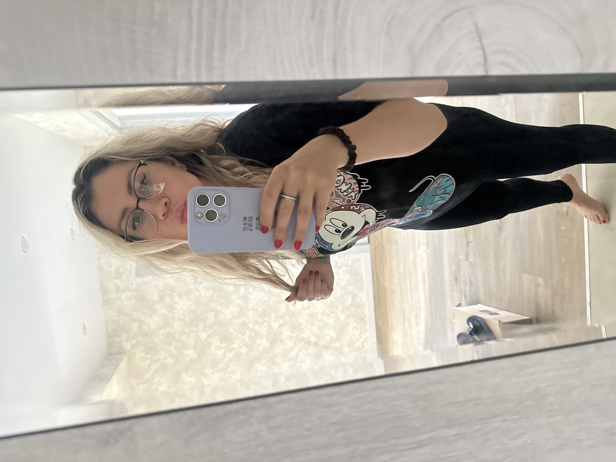 A woman takes a mirror selfie in a room with light-colored patterned wallpaper and wooden floors. She is wearing black leggings, a black t-shirt with a Mickey Mouse graphic, glasses, and a silver ring. Her hair is blonde and wavy, and her fingernails are painted red. She is holding a light purple phone and has one hand in her hair. In the reflection of the mirror, a cardboard box with a pair of black shoes is visible on the floor. The scene is located in Novoaltaysk, Russia.