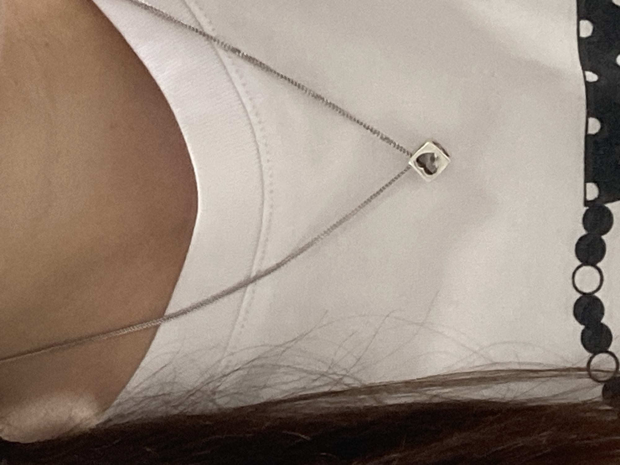 The image shows a close-up of a person wearing a white t-shirt. A delicate silver necklace with a square pendant containing a heart-shaped cutout is visible, resting on the t-shirt. The person's neck and chest area are the primary focus. The background is out of focus and appears to be an indoor setting. There is no indication of the city of Poltava, Ukraine, or any outdoor environment.
