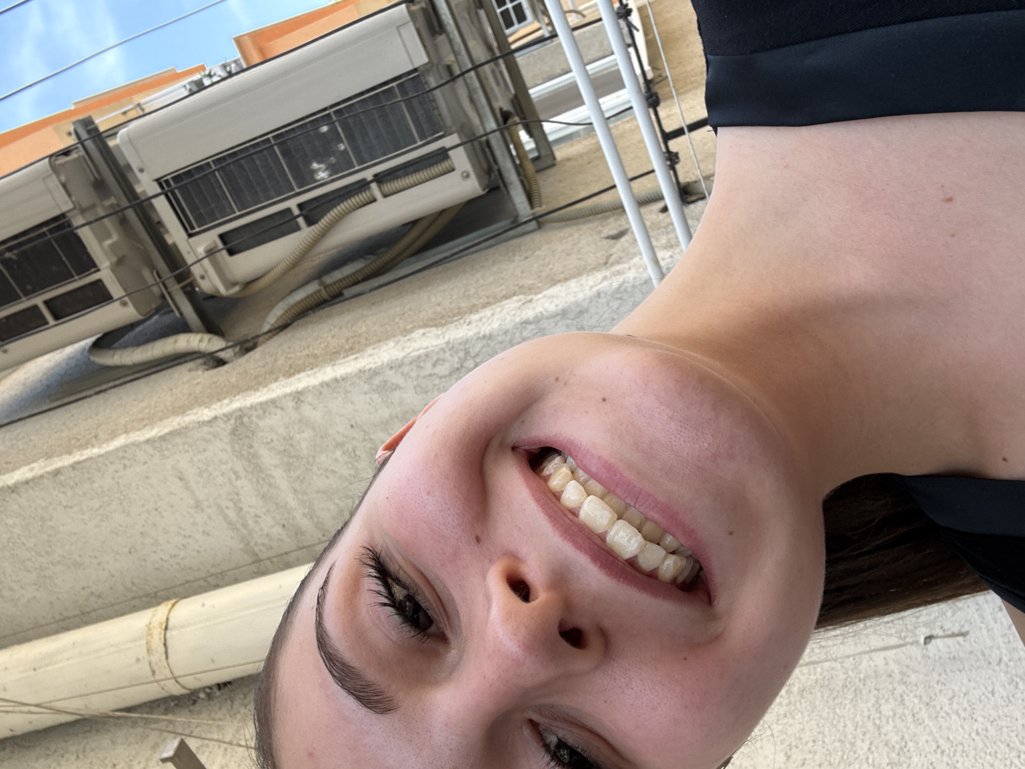 This close-up selfie-style image is rotated 90 degrees clockwise, featuring the smiling face of a young woman occupying the lower right half of the frame. Her head is tilted, and she is looking upwards relative to the background. She has dark hair, dark eyebrows, and long eyelashes. Her expression is one of broad happiness, with a wide smile revealing her natural teeth. A black top or garment is visible on her shoulder and neck area.
The background, appearing tilted upwards due to the image rotation, shows an urban setting. In the top left, a bright blue sky is visible, indicating daytime. Below the sky, an orange-colored building or structure can be seen. The majority of the background consists of a textured, light-colored concrete wall or building exterior, where several outdoor air conditioning units are mounted. These units, along with visible wires, pipes, and some white vertical poles, suggest an older residential or commercial building. One pipe appears dirty with a rust-like stain.
Given the rotation, the woman appears to be holding the camera and looking upwards at the building facade. The clear blue sky indicates sunny weather. The location is specified as Burgas, Bulgaria, consistent with such building exteriors featuring external AC units. There is no visible text in the image.