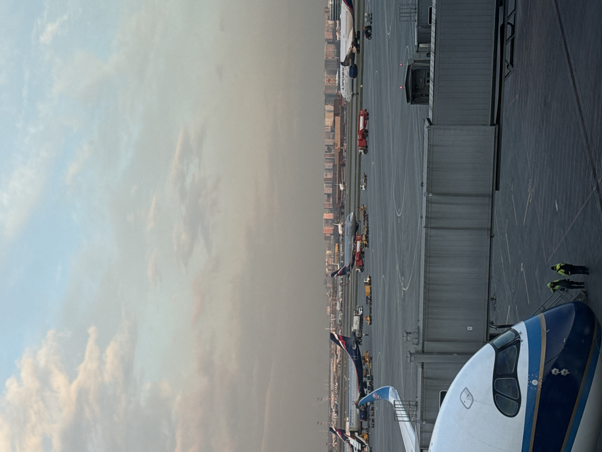 The image, when viewed in its correct orientation, captures a wide view of an airport tarmac under a hazy, partially cloudy sky, suggesting either early morning or late afternoon light. In the foreground, the distinctive blue and white nose of a passenger aircraft is prominently visible, with two airport ground crew members in high-visibility yellow-green vests standing near its front.
The tarmac is active with several other airplanes parked at gates. One large white aircraft clearly displays "АЭРОФЛОТ" (AEROFLOT) in Cyrillic lettering on its fuselage, identifiable by its red and blue tail. Various ground support equipment, including red baggage tugs and service vehicles, are scattered across the apron. Multiple jet bridges are connected to parked aircraft or positioned nearby, indicating ongoing boarding or deplaning procedures.
In the background, a distant city skyline is visible beneath the muted sky, which transitions from a lighter blue with visible clouds on the left to a more uniform, hazy grey on the right. The scene depicts typical operations at a busy international airport. The presence of Aeroflot aircraft and the location context suggest this is Sheremetyevo International Airport near Khimki, Russia.