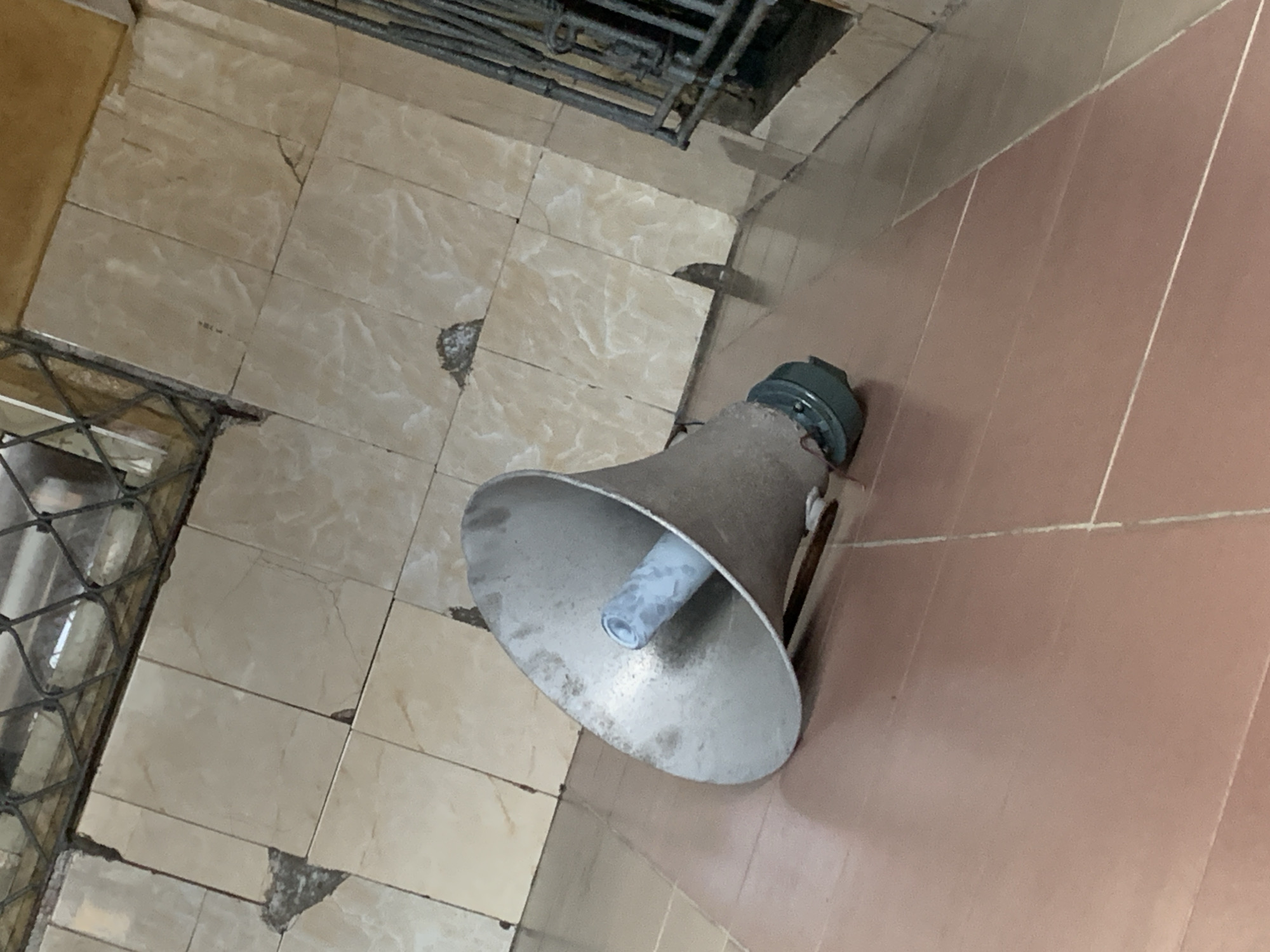 A wide-angle, slightly tilted, medium shot captures an old, metallic megaphone lying on a tiled floor in Alaiyabiagba, Nigeria. The megaphone, which appears to be weathered and somewhat dirty, is positioned on its side, with its wide bell facing the viewer and its narrower end pointing away. The bell has a dusty, matte silver finish, and a light blue cylindrical component is visible inside the narrow opening. The megaphone is resting on a floor covered in large, rectangular, reddish-brown tiles. Behind the megaphone, the floor transitions to a different set of larger, beige tiles that are cracked and chipped in several places, suggesting disrepair. A metal grate, likely part of a fence or window, is visible in the upper right and upper left corners of the image, adding to the sense of a neglected urban environment. The overall lighting is dim, contributing to the somber atmosphere of the scene. There are no people or animals visible in the image, and no discernible actions are taking place. The environment appears to be an outdoor or semi-outdoor space, possibly a public area or a forgotten corner of a building.
