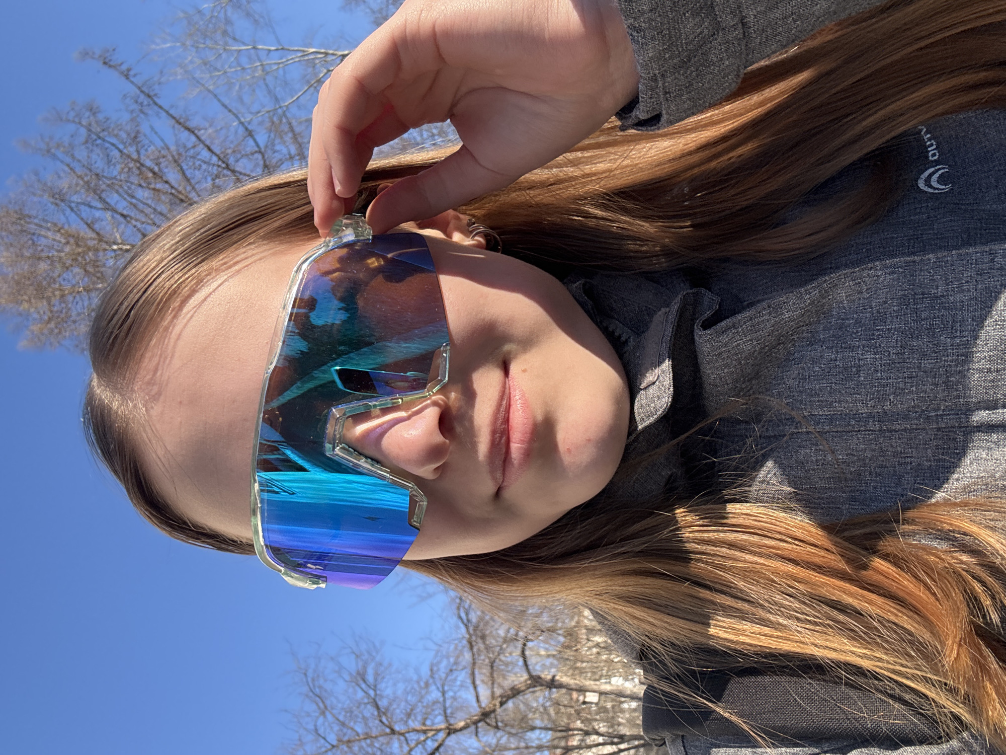 A young person with long, reddish-brown hair is outdoors, tilting their head to the side and holding futuristic-looking iridescent blue and silver sunglasses. The person is wearing a gray jacket with a logo visible near the collar that says "OUTV". The background shows a clear blue sky with bare tree branches. The location is potentially a sunny area in Berezniki, Russia, based on the reference, although specific landmarks are not visible.