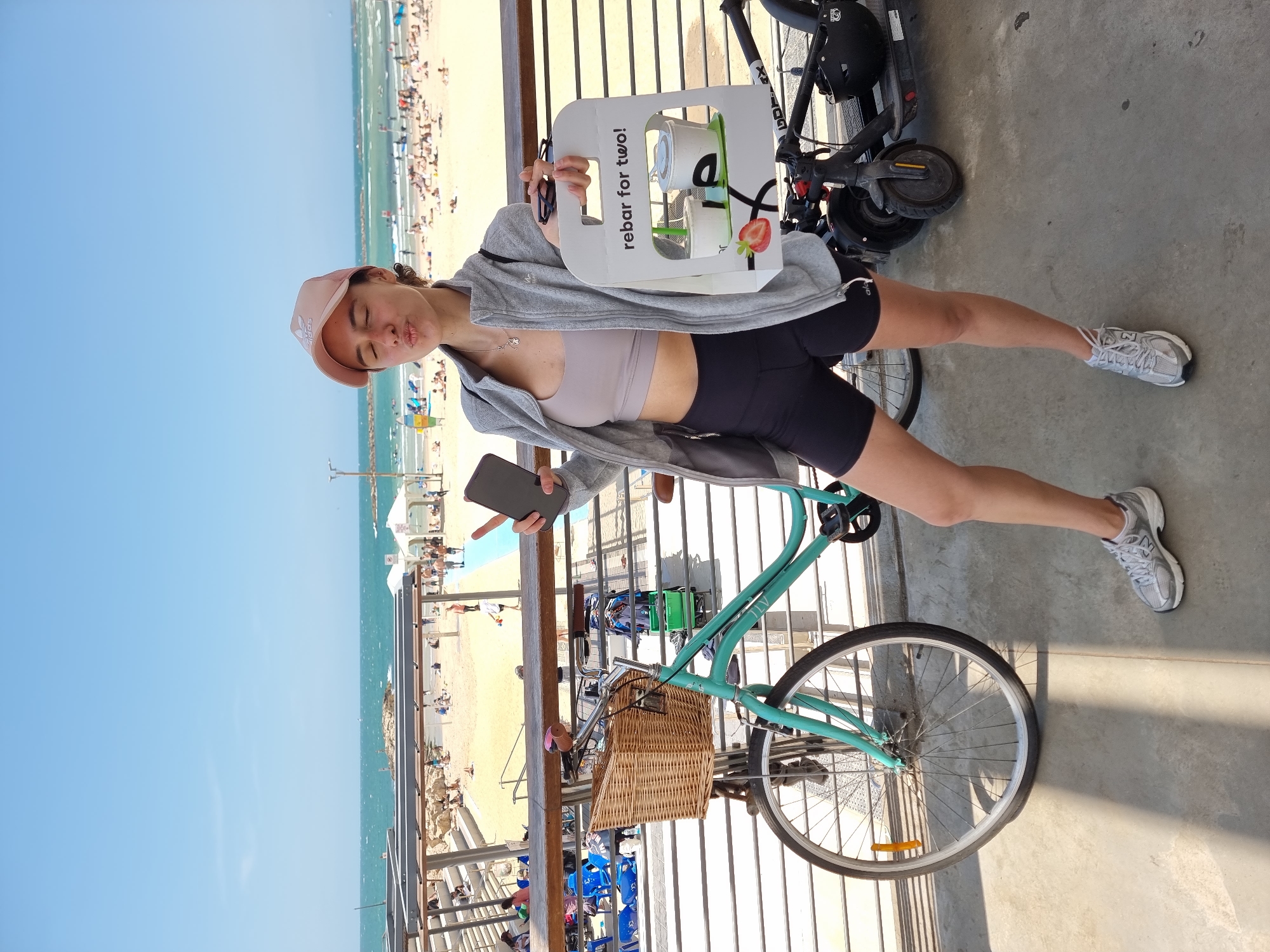 The image shows a woman on a beach promenade. She is wearing a pink baseball cap, a light grey hooded jacket, a beige sports bra, and black shorts. She is holding a smartphone in one hand and a "rebar for two!" container in the other, with sunglasses perched on top. The container appears to contain two cups of a drink with a strawberry on the side.
A turquoise bicycle with a wicker basket is beside her, and a black electric scooter is parked nearby. The background shows a sandy beach filled with people, the Mediterranean Sea, and a clear blue sky. The scene appears to be taken along the Tel Aviv beach promenade in Israel.