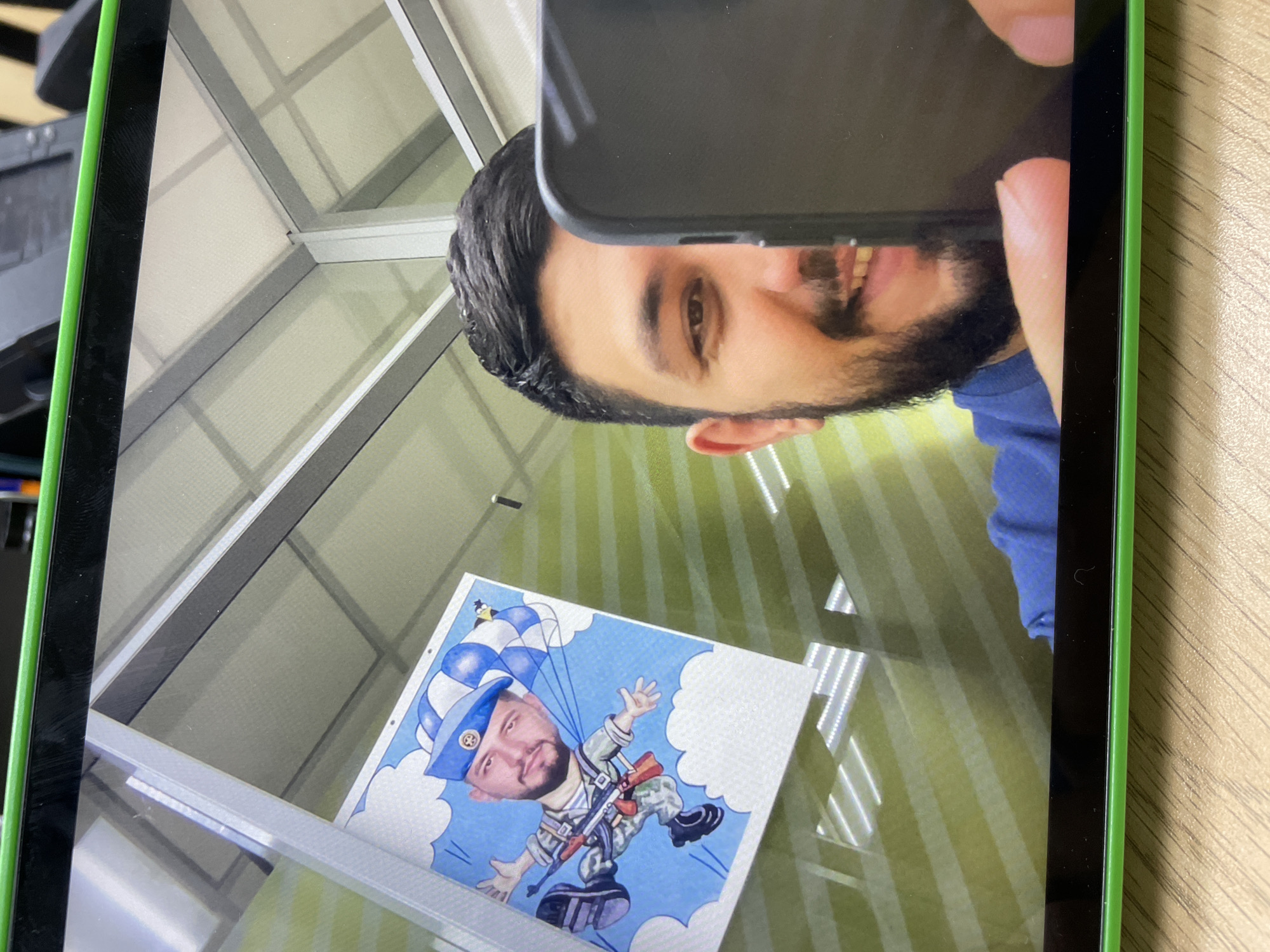 A photograph displays a man holding a mobile phone, angled to capture his face. The phone obscures the upper portion of his face, but the image reveals his dark hair, beard, and a portion of his smile. The background reveals a structured ceiling with a pattern of beams and panels. Below, a printed caricature shows a figure in military attire with a parachute, seemingly based on the man's likeness. This scene is displayed on a tablet with a green border. Based on the caricature's uniform and the likely context, the location could be in Krasnoyarsk, Russia, referencing Russian Airborne Troops. The photograph captures the act of taking a picture of the tablet.
