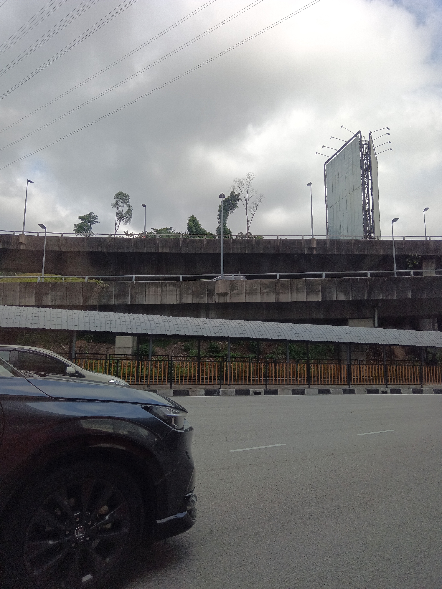 The image shows a street scene in Kuala Lumpur, Malaysia. In the foreground, the front left quarter of a dark-colored car is visible, driving on an asphalt road with white lane markings. In the background, a multi-level overpass is prominent, with concrete structures and railings. A pedestrian walkway with a grey, corrugated roof runs beneath the overpass. Beyond the overpass, a tall, rectangular billboard structure stands against a cloudy sky. Several streetlights and electrical wires are also visible throughout the scene, and scattered trees are present along the top of the overpass. The overall environment suggests an urban setting with infrastructure for transportation and advertising.