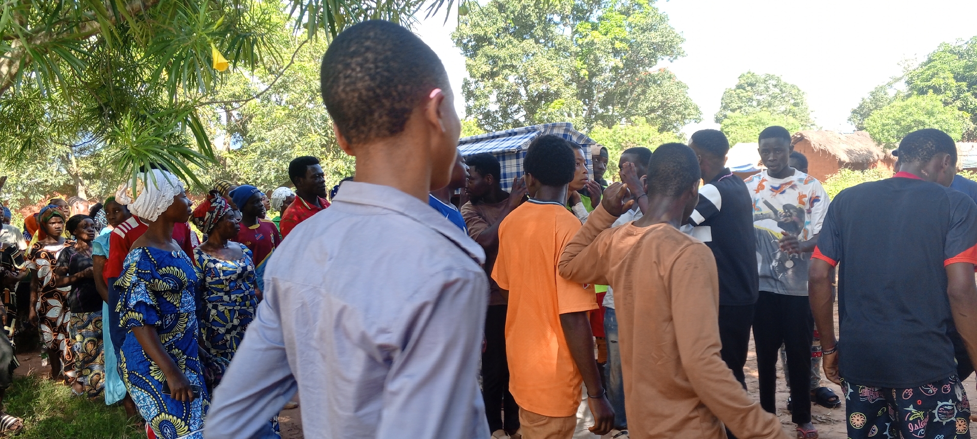 A daytime shot captures a group of people gathered in Ngalula, Democratic Republic of the Congo. The setting is outdoors, surrounded by trees and under sunlight, where a group of people stand around.
On the left, a line of women stand out in colorful patterned dresses. In front, a man with a gray shirt is facing away from the camera. A separate cluster of young men in casual attire, like t-shirts, stand closely, facing the right side of the frame. In the background, a house with a thatched roof, partially visible.