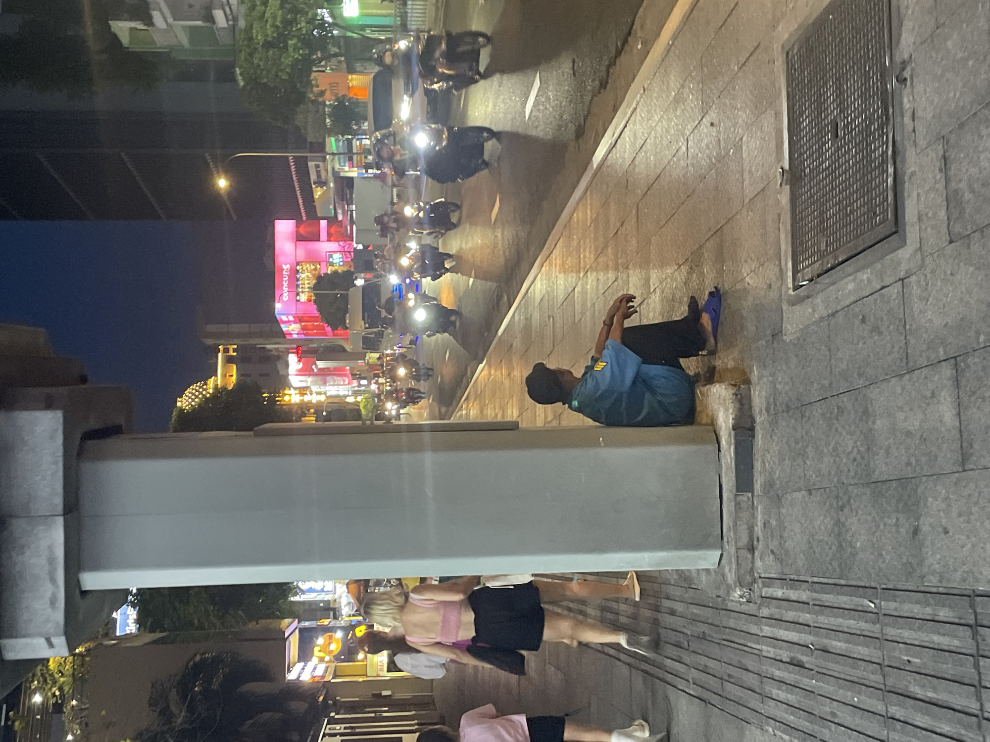 A sidewalk in Thủ Đức City, Vietnam, is shown at night. A man wearing a blue uniform and a dark cap sits on the ground next to a concrete pillar. He is looking down at his hands, which are resting on his knees. In front of him is a metal grate. To his left, a woman with blonde hair wearing a pink crop top and black shorts walks away from the camera, followed by a younger person wearing a pink t-shirt. Across the street, traffic, primarily motorcycles, moves along the road, their headlights illuminating the scene. Buildings and illuminated signs are visible in the background.