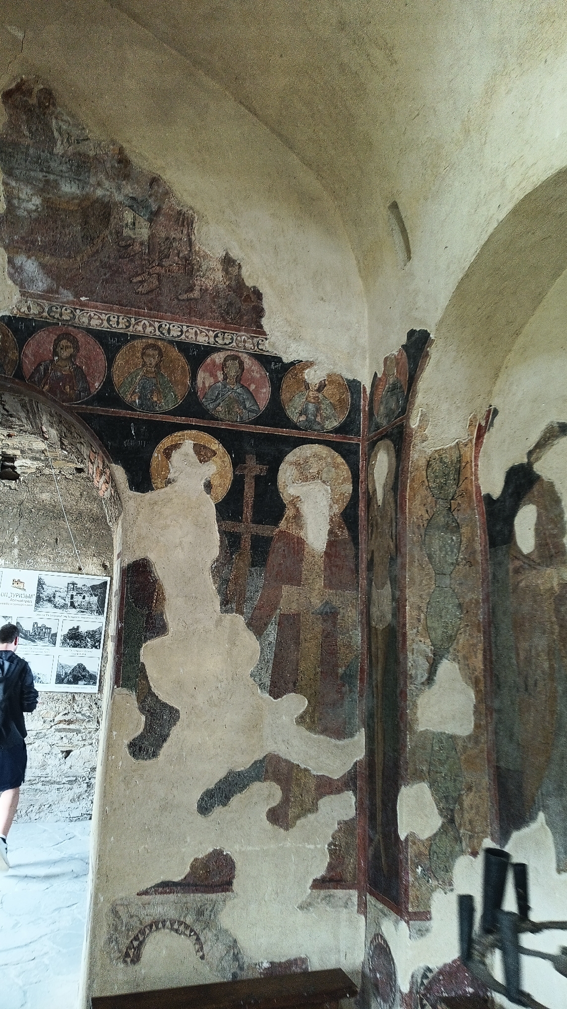An interior view reveals a partially deteriorated mural within a stone structure. The mural features depictions of figures with halos, religious iconography like crosses, and circular portraits above. The paint is peeling and damaged, exposing the underlying wall in large sections. A person wearing a black jacket is walking away from the camera in the background. A series of black and white images are posted on a board beside the person. The architectural style is old and worn, with visible deterioration and age. A metal railing stands near the wall at the lower right. The city of Unknown, Unknown is not known.