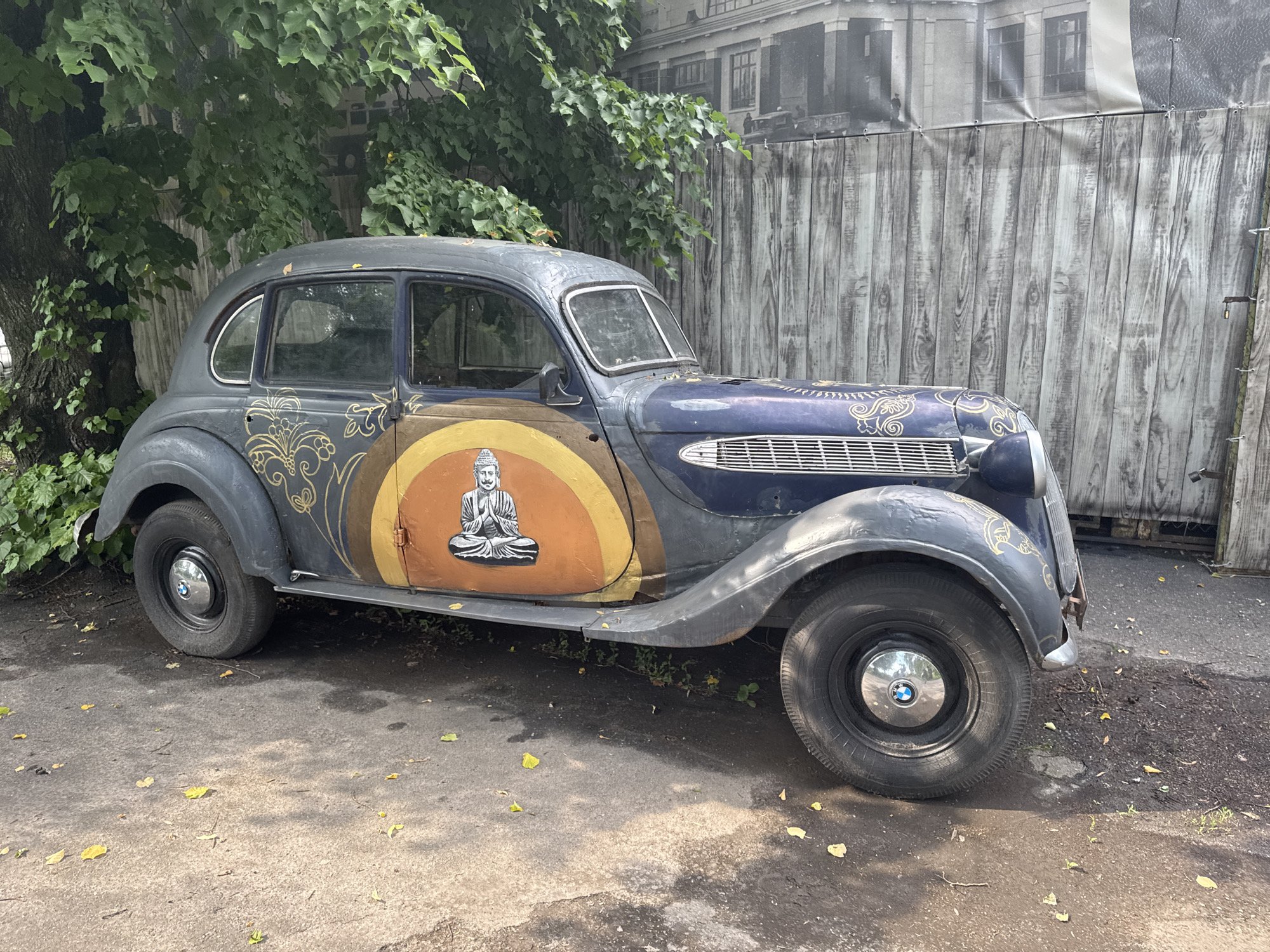 An antique dark blue BMW sedan, featuring a painted orange and yellow circular design on the front driver's side door with a meditating Buddha figure, is parked outdoors on a paved surface, partially shaded by a large tree with green leaves. Behind the car, a wooden fence-like structure serves as a backdrop, with a large banner depicting a building facade hanging above it. The ground is scattered with fallen yellow leaves and small green plants. The scene appears to be a static display, possibly in a public space or exhibition area within the city of Vinnytsia, Ukraine.