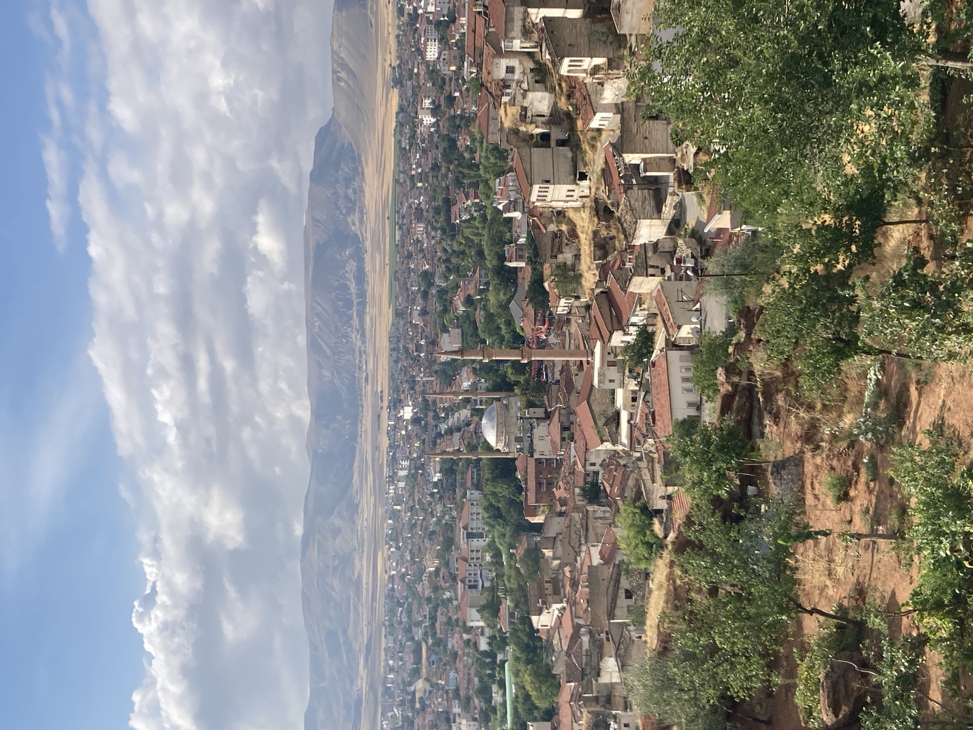 The image captures a panoramic view of Avanos İlçe Merkezi, Turkey, on a partly cloudy day. In the foreground, dry, rocky terrain with sparse vegetation and a few trees slopes downwards. The midground reveals a densely populated urban area with a mix of old and new buildings. Many structures have red tile roofs, characteristic of older architecture, interspersed with modern buildings. Prominently visible are two mosques with tall, slender minarets and domed roofs, suggesting the religious and architectural identity of the area. The background features a vast, arid landscape with gently rolling hills and mountains under a sky filled with large, puffy white clouds. The overall impression is one of a historic town nestled within a rugged, natural environment.