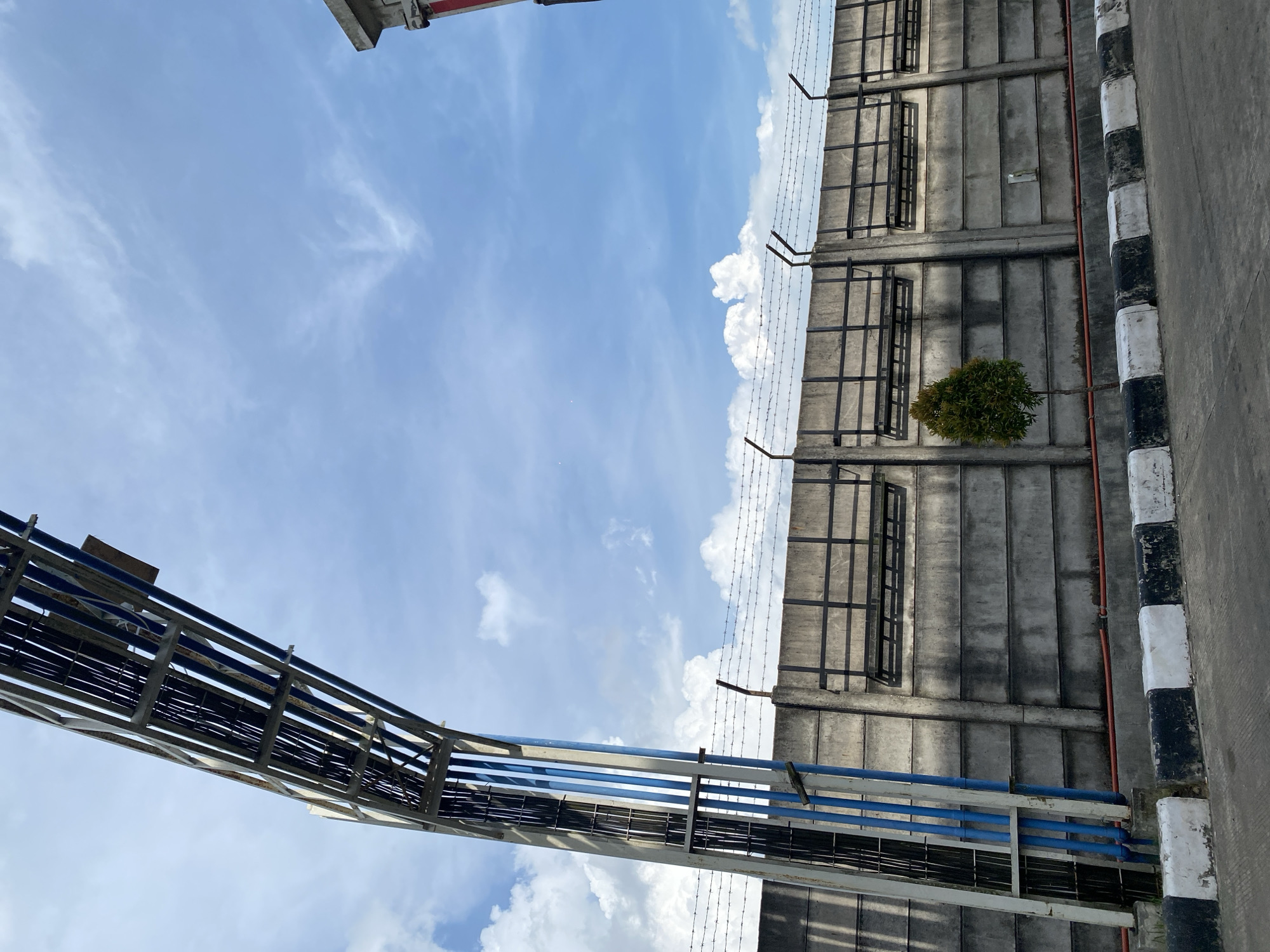 In Gambut Barat, Indonesia, a concrete wall topped with barbed wire stretches across the frame. Above the wall, a large metal structure supports numerous cables and pipes, extending from the left side of the image into the sky. The sky is a vivid blue with scattered white clouds. In front of the wall, a small, green tree stands, and a narrow red pipe runs along the base of the wall. The foreground features a paved surface with a curb marked by alternating black and white blocks. The scene appears to be outdoors, possibly in an industrial or utility area.