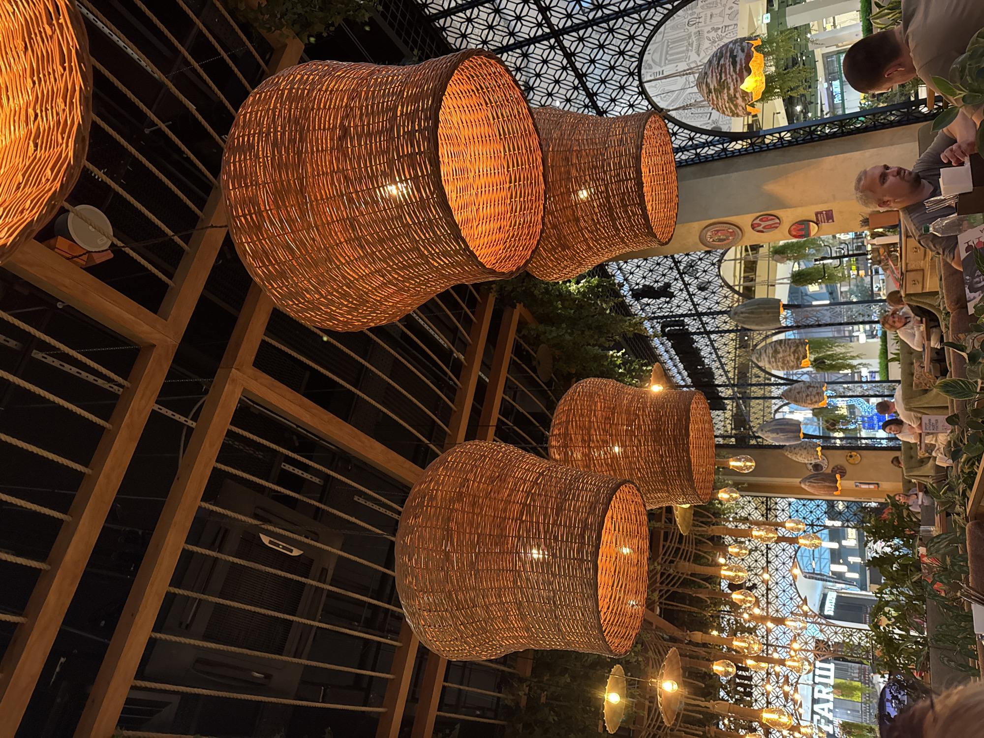 The image displays an indoor restaurant or cafe setting within a multi-story building, likely a shopping mall or large atrium, in Perm, Russia, during the evening or night. The primary subjects are numerous large, cylindrical pendant lamps crafted from woven wicker, which emit a warm, orange light, creating a cozy ambiance. These lamps are suspended from a decorative ceiling structure made of wooden beams and ropes.
Several people are visible, primarily seated at tables on the right side of the frame, engaged in dining or conversation. One man with short hair and a beard looks towards the left side of the image, while another man is partially visible further in the background. Other patrons are discernible in the distance. The decor includes potted greenery and additional decorative string lights with exposed filament bulbs.
Notable details include the intricate patterns of the upper ceiling and reflections on glass surfaces, indicating a spacious interior. The lighting suggests it is past daylight hours. Visible text includes a sign reading "FARШ" in Cyrillic, which translates to "FARSH," with "BURGER & GRILL" beneath it, identifying a food establishment. The overall scene conveys a relaxed and contemporary dining atmosphere.