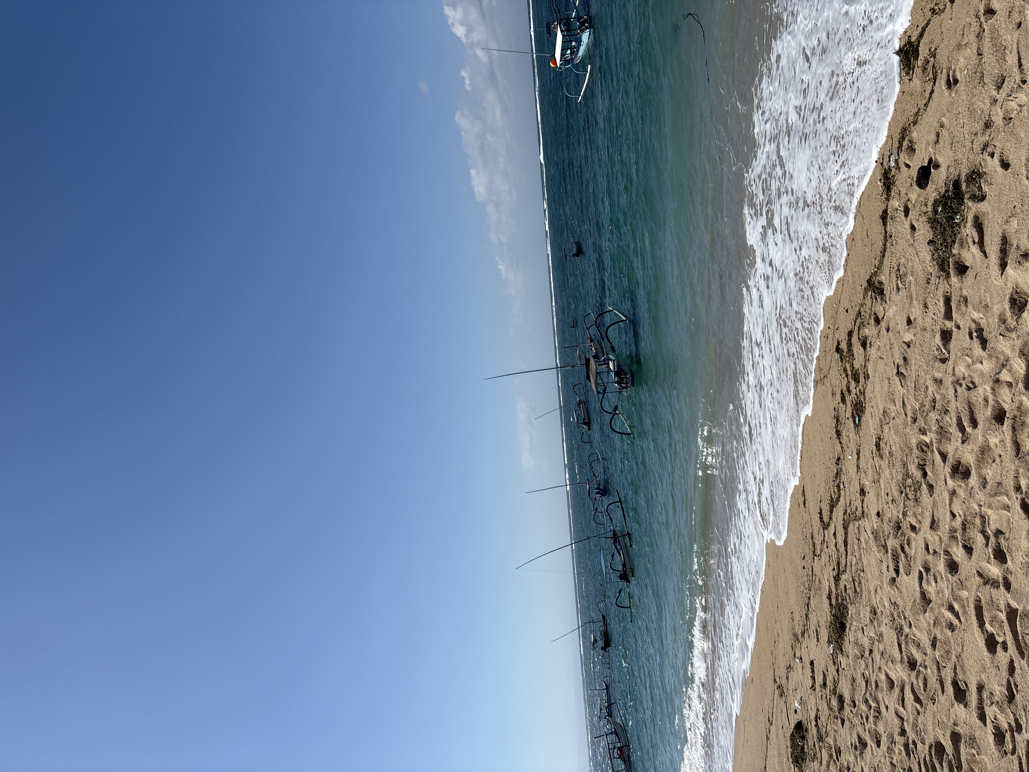 The image displays a vibrant, sunny beach scene, characteristic of a tropical location like Kuta Selatan, Indonesia. The upper left portion of the frame features a clear, bright blue sky with only a few faint, wispy white clouds.
Horizontally across the middle, the serene, turquoise-to-teal colored ocean extends. Several traditional Indonesian outrigger boats, known as Jukungs, are visible floating in the water. These boats, with their distinctive outriggers, appear to be either moored or gently bobbing in the calm conditions.
On the right side of the image, the golden-brown sandy beach curves, meeting the ocean where gentle, foamy white waves are breaking softly. The sand shows numerous footprints, indicating recent human presence, and scattered patches of dark green seaweed or debris are visible along the shoreline.
The scene conveys a peaceful, relaxed atmosphere under bright sunshine, suggesting it is daytime, likely morning or early afternoon, with clear weather. There is no visible text in the image.