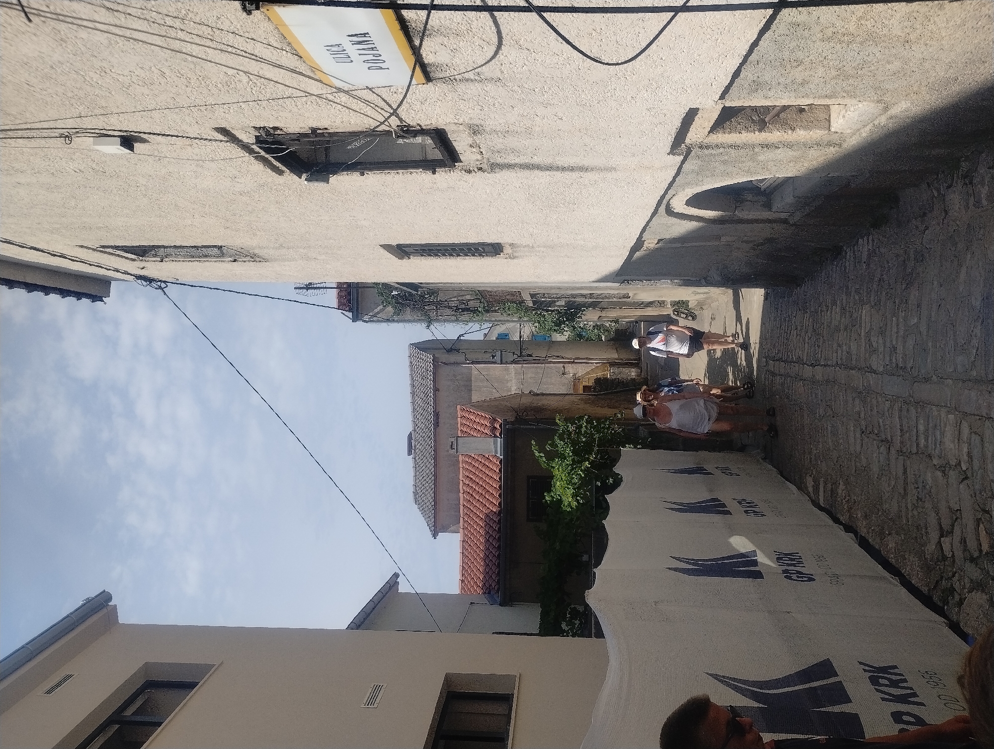 In Općina Vrbnik, Croatia, a narrow cobblestone street is bordered by buildings. On the right, a pale beige building features a sign that reads "ULICA POJANA". Several people are walking away from the camera down the street. A white tarp with blue lettering and logos is installed along the left side of the street, partially obscuring the view of the buildings behind it. The sky is cloudy.