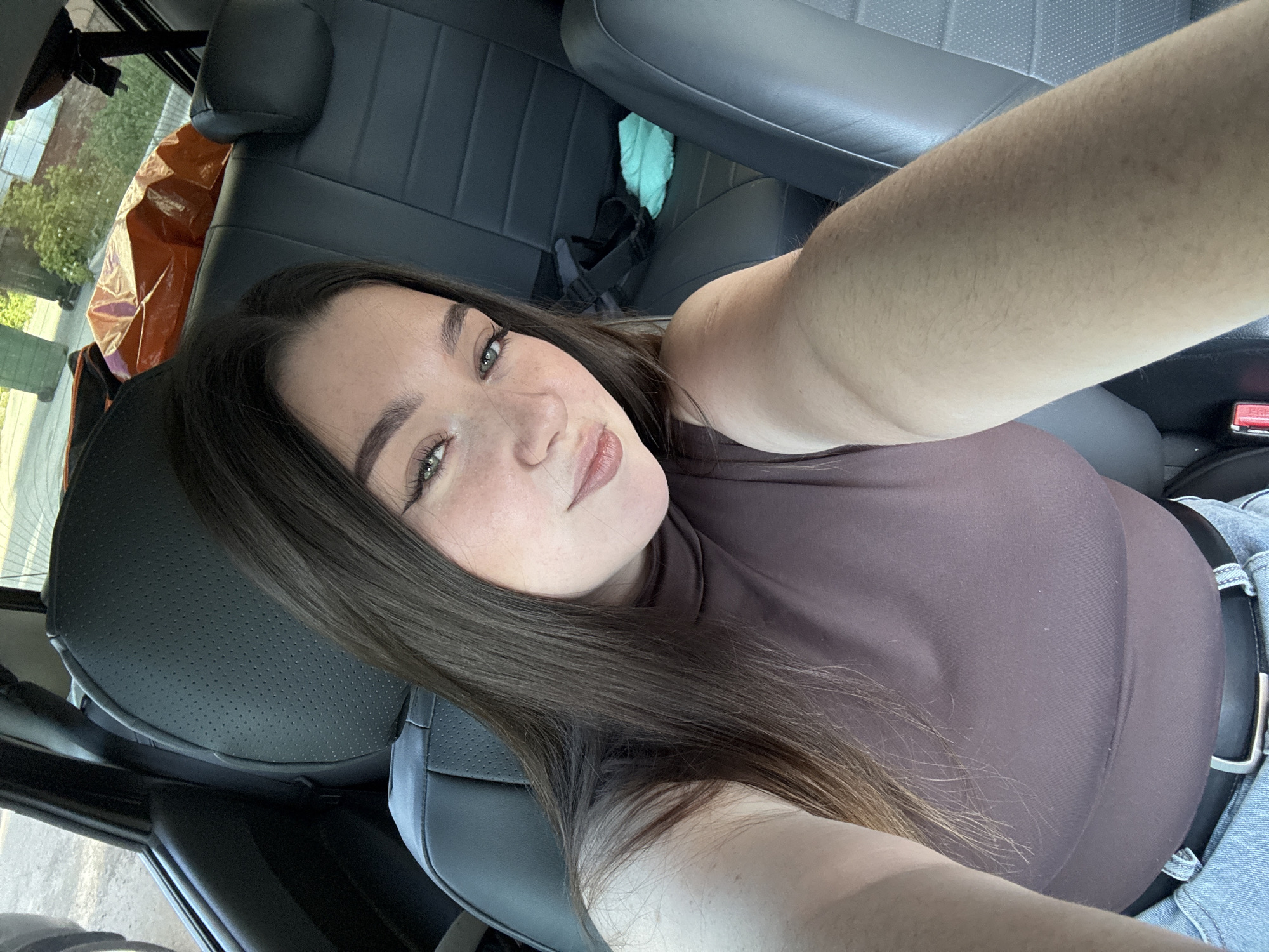 A woman with long brown hair and fair skin takes a selfie in what appears to be the driver's seat of a car. She's wearing a brown top. The car's interior is visible, including the seat and dashboard. Through the window, a building and some greenery can be seen. No definitive details indicate the location is Perm, Russia.