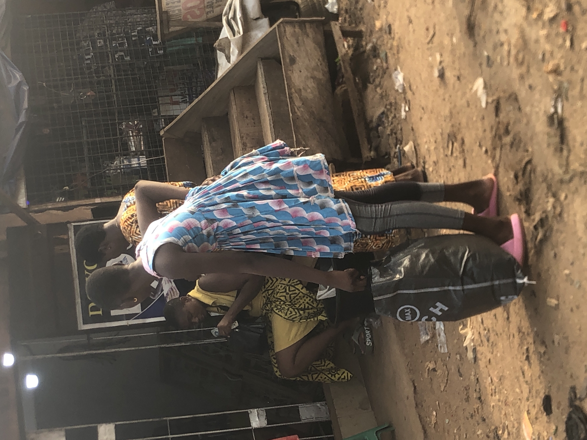 This daytime image, taken in Onitsha, Nigeria, captures a bustling outdoor market or street scene. The ground is unpaved, dusty, and uneven, with scattered debris, suggesting a busy urban environment.
In the foreground, a woman stands centrally on the left, wearing a blue and white patterned, pleated dress over dark leggings, complemented by bright pink flat shoes or slippers. She is looking downwards and slightly to her right. To her immediate right, a large black bag, possibly a duffel or storage bag, rests on the ground. White lettering on the bag clearly displays "MAX" and "SPORT."
Behind the central figure, another woman is partially visible, dressed in an orange and brown patterned garment. Further right and lower to the ground, a child in a yellow patterned outfit is seen sitting or crouching. Another child's head is visible just to the right of the child in yellow. The individuals appear to be congregated, possibly engaging in commerce or social interaction typical of a market setting.
The background reveals simple commercial structures: a dark metal grid or fence-like barrier and wooden shelves or stairs. A partially visible sign behind the people shows the letters "DVC" in white text on a dark background. Other faint text, including what appears to be "20", is present on a lighter sign in the upper right portion of the background. The bright, ambient lighting confirms it is daytime. The scene conveys a sense of everyday life and activity in the Nigerian context.