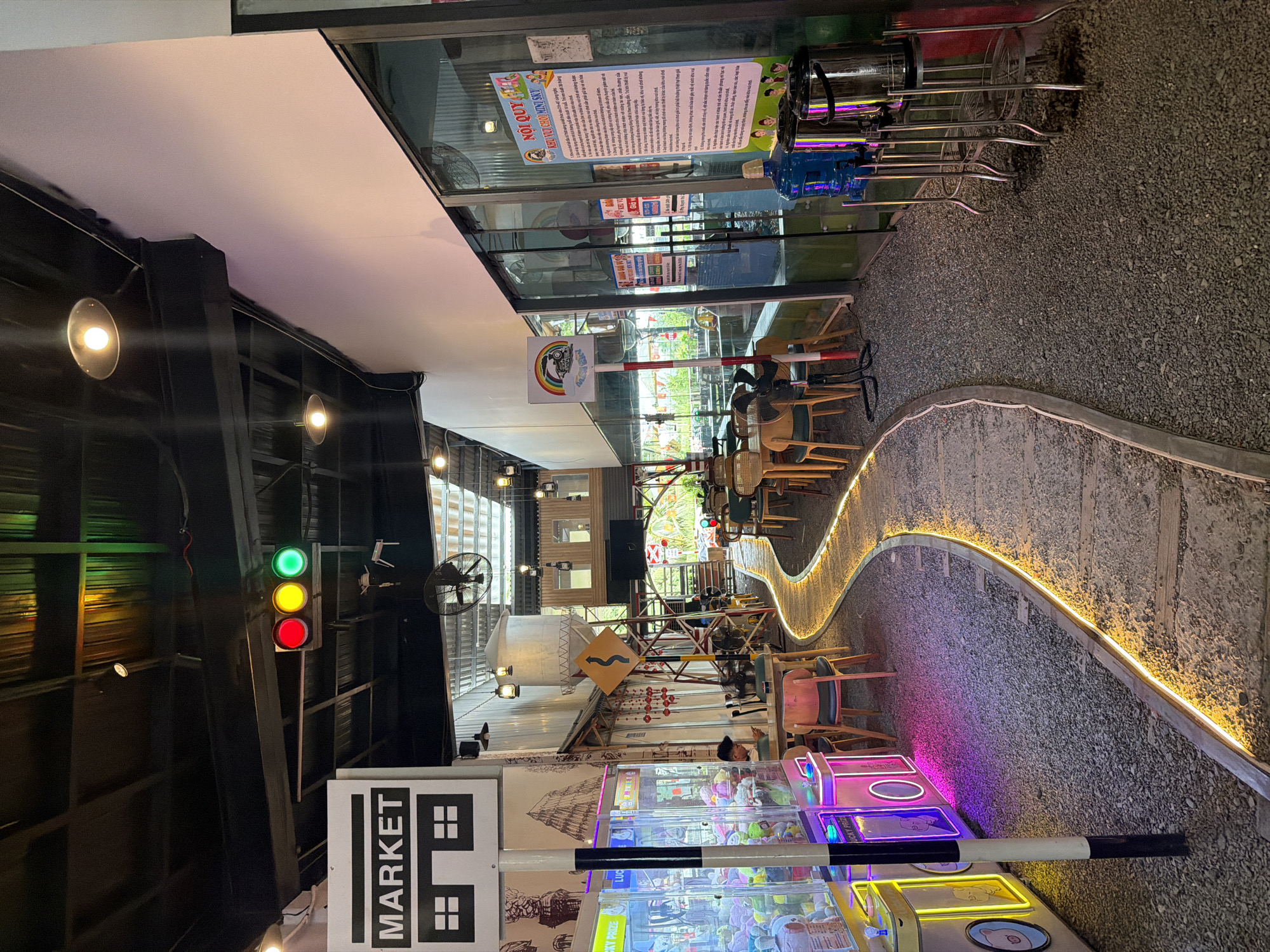 This is an indoor recreation area with elements of a marketplace, likely located in Nha Trang Ward, Vietnam, as indicated by the Vietnamese text on a sign. The scene appears to be arranged with a decorative, winding pathway illuminated with yellow lights, bordered by gravel.
On one side of the pathway, there are tables and chairs suggesting a dining or seating area. Further into the space, there are claw machine arcade games lit up with neon pink and yellow lights.
In the foreground, a sign reads "MARKET" with a graphic of a building with windows. Next to it stands a black and white striped barrier. A traffic light is also visible.
The environment consists of a combination of open-air and enclosed structures, with natural light filtering in through open areas. The space is adorned with various themed decorations, including road signs. The lighting includes overhead lamps and neon lights.