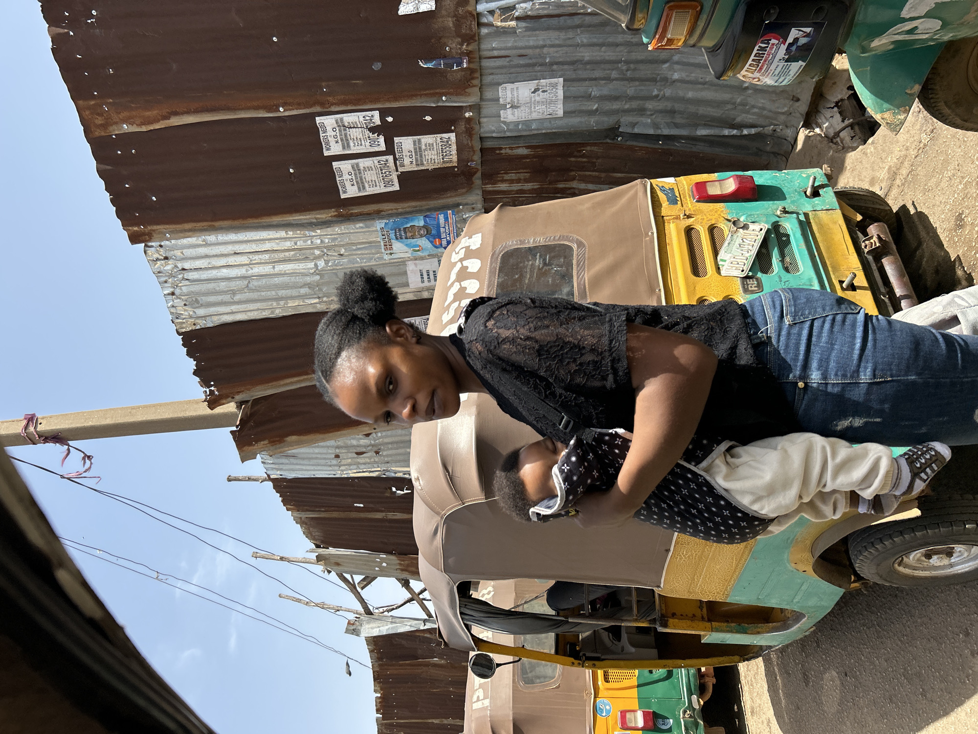 A medium eye-level shot depicts a woman holding a baby in front of a three-wheeled motor vehicle (auto rickshaw) against a backdrop of corrugated metal structures and a partially visible cityscape.
The woman, dressed in a black lace top and blue jeans, cradles the infant, who is wearing a patterned wrap and light-colored clothing. The auto rickshaw, which features a faded yellow, green, and brown color scheme, bears a license plate and an advertisement sticker.
The location is suggested to be Jos, Nigeria, based on the presence of an auto rickshaw and an advertisement sticker. The surrounding corrugated metal structures and posters attached to the walls contribute to the depiction of a street scene.