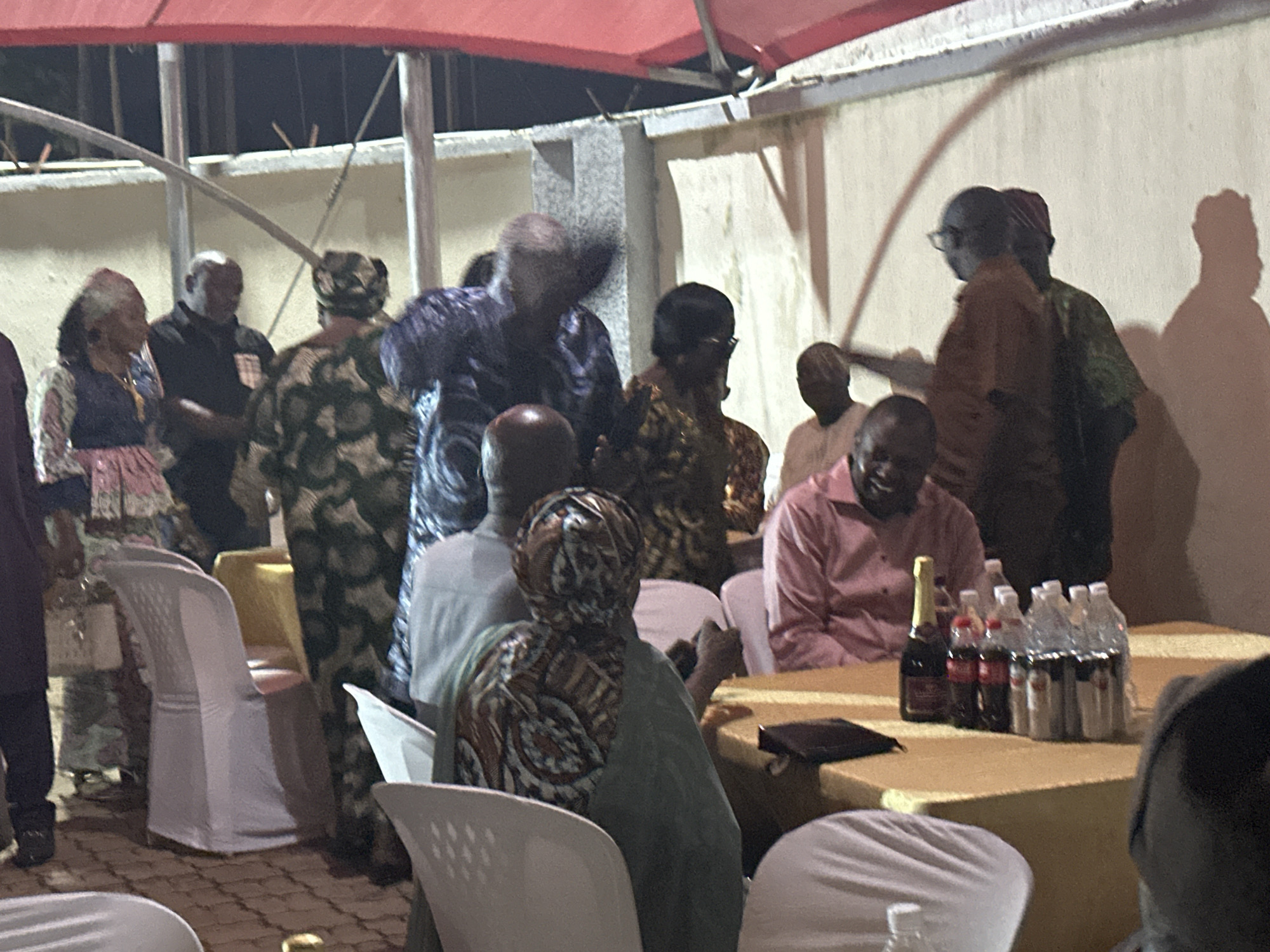 A group of people are gathered outdoors in what appears to be a social event. The location is Narayi, Nigeria. Individuals are seen standing and sitting around tables, dressed in various styles of clothing. Several water bottles, soda, and a bottle of champagne are on the table. The setting has the appearance of a celebration or gathering.