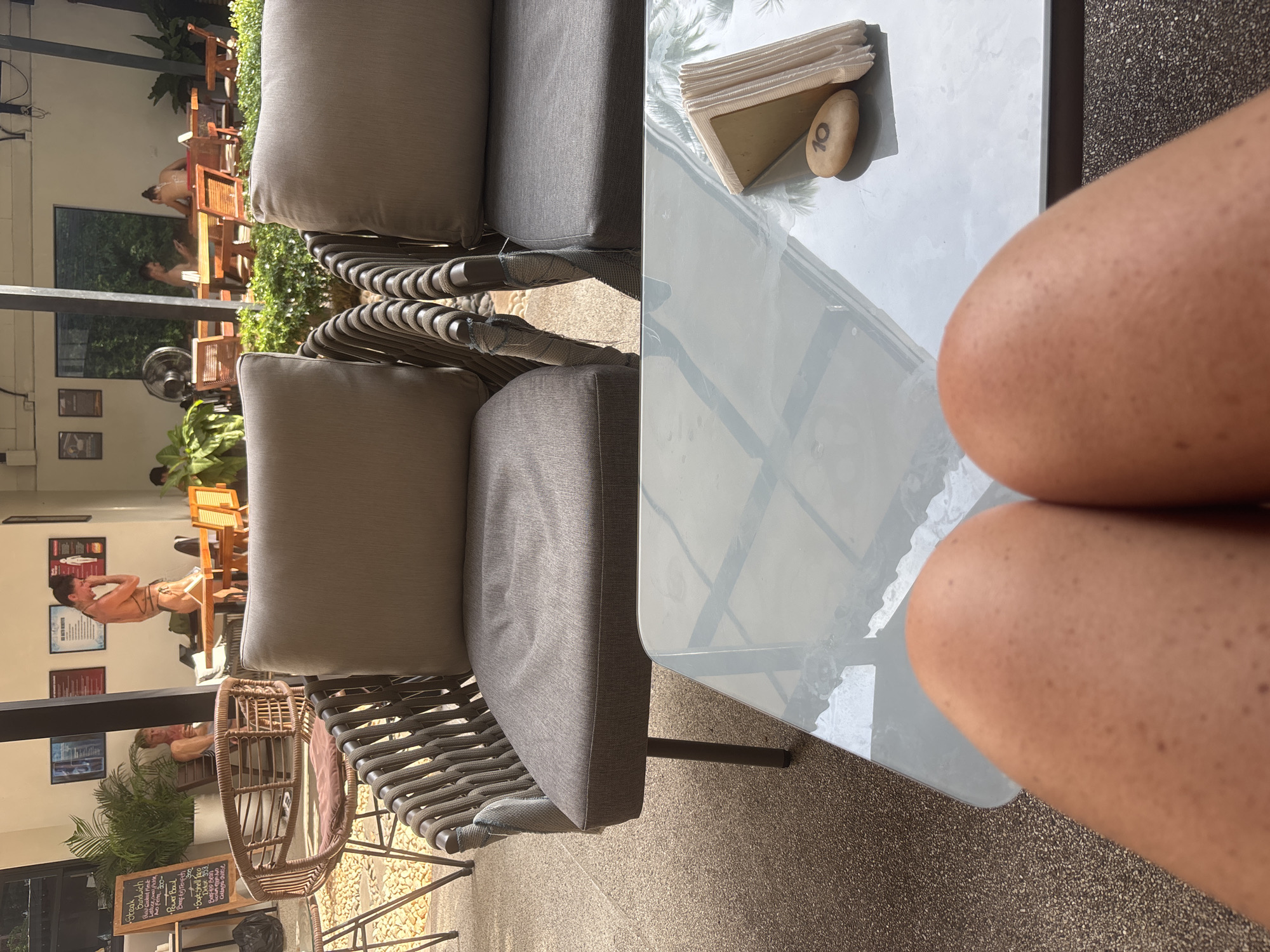 In a cafe setting in Ban Rak, Thailand, an outdoor seating area features two plush grey armchairs and a glass-topped table. The table holds a napkin holder, a small pebble with the number "10" written on it, and is partially obscured by a person's legs, suggesting the image is taken from their perspective. In the background, several people are visible at tables and walking around. One woman in a bikini stands by a table, while others are seated, some with phones in hand. Lush green plants are strategically placed throughout the outdoor space, and a black fan is visible. A blackboard menu is propped up near one of the chairs. The lighting suggests it is daytime.
