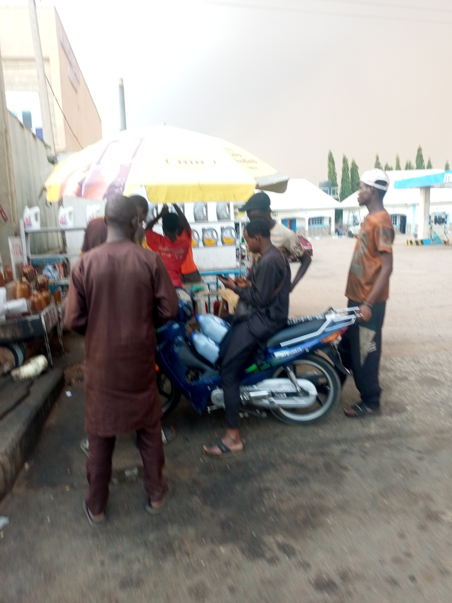 The image depicts a scene at a roadside petrol station in Gombe, Nigeria. Several men are gathered around a blue motorcycle. One man is sitting on the motorcycle, looking at a phone. Other men are standing nearby, and a few are near the front of the motorcycle. Behind them is a shaded area with motor oils and oil bottles. In the background, there's a building. The sky appears overcast.