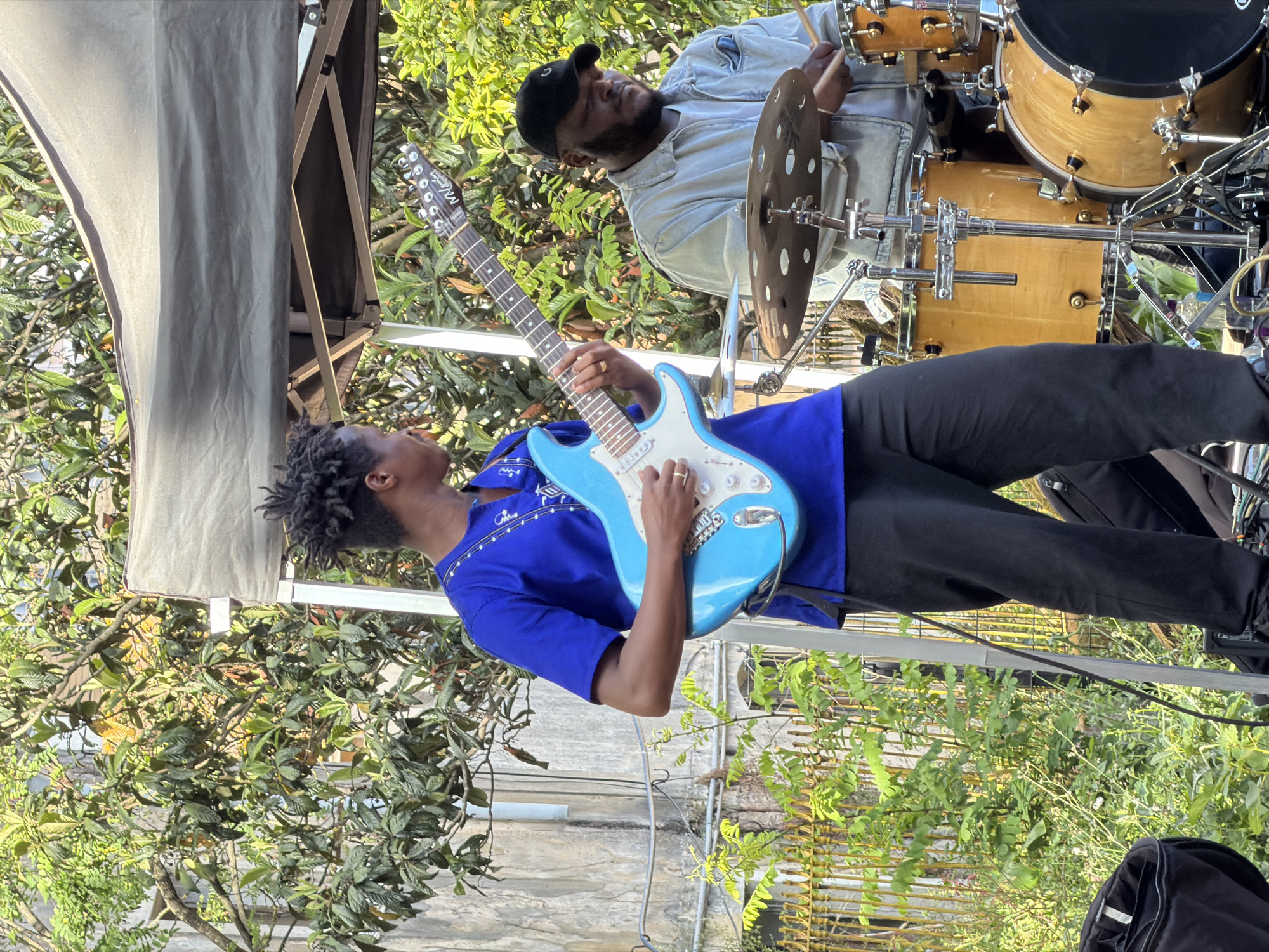 A medium shot shows a guitarist and drummer performing outdoors, in what appears to be Lisbon, Portugal. The guitarist, with dreadlocks and a royal blue shirt with white embellishments, plays a light blue electric guitar. To his right, a drummer wearing a denim-like shirt and a black baseball cap is behind a drum set. A dark canopy is visible in the upper left, and the background features lush green foliage, suggesting a park or garden setting. A weathered stone wall is partially visible in the lower left. Wires hang in the scene. In the lower right is a black object that appears to be a bag.