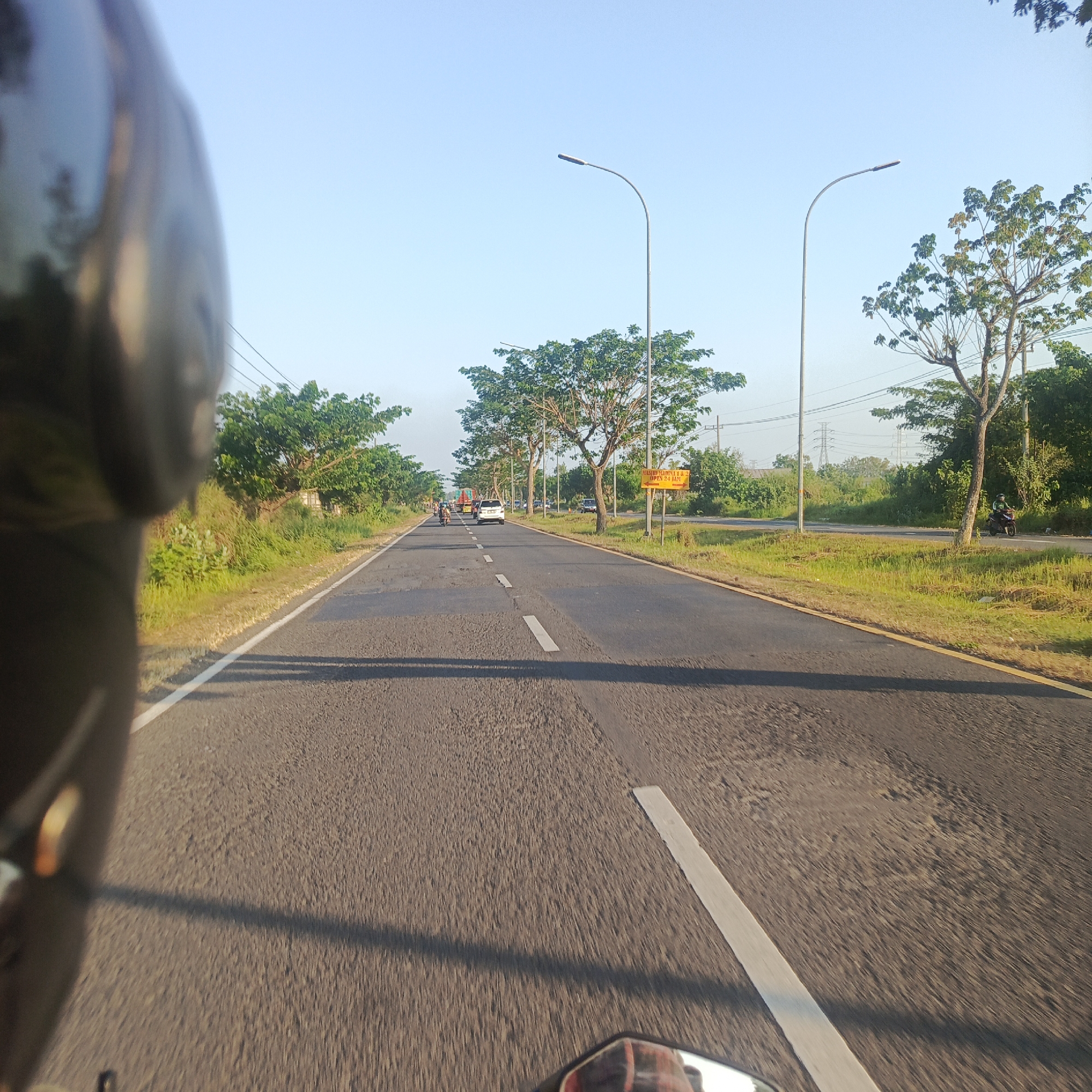The image depicts a view from a motorcycle traveling along a straight, paved road with a yellow line and a white broken line marking the lanes. The road is flanked by grassy verges and lined with mature trees. In the distance, several vehicles, including cars and trucks, are visible on the road. The sky is clear and blue, suggesting it is daytime. On the right side of the road, a yellow sign is posted with text that appears to be in Indonesian. This scene is located in Petapan, Indonesia.