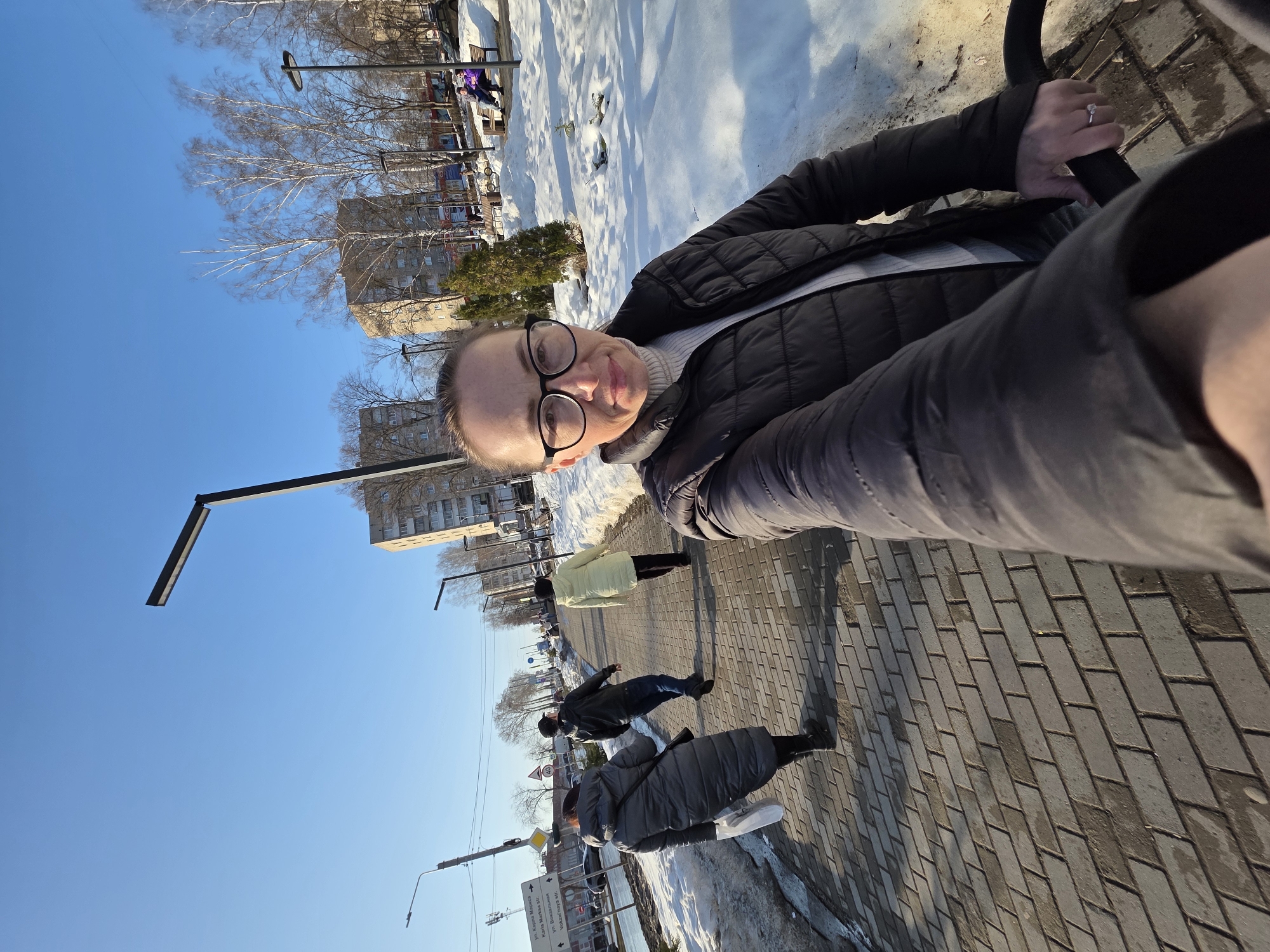 An outdoor wide-angle selfie depicts a Caucasian female, mid-frame right, wearing black-rimmed eyeglasses and a black puffer jacket over a light turtleneck. Her right hand, adorned with a ring on the left ring finger, grasps a black handle. She is positioned on a grey brick-paved sidewalk adjacent to a snowy park area. A street sign in the background identifies the location as the intersection or vicinity of Karl Marx Street ("ул. Карла Маркса") and Vokzalnaya Street ("ул. Вокзальная") in Chaikovsky, Russia. Three individuals walk away from the camera on the sidewalk: a female in a dark grey puffer coat carrying a white bag, a male in a black jacket and cap, and a female in a light yellow puffer coat. In the snowy park area to the right, two individuals are seated on a wooden bench. Multiple modern black streetlights and multi-story buildings are visible under a clear blue sky. Patches of melting snow are present on the ground, indicative of a winter or early spring environment.