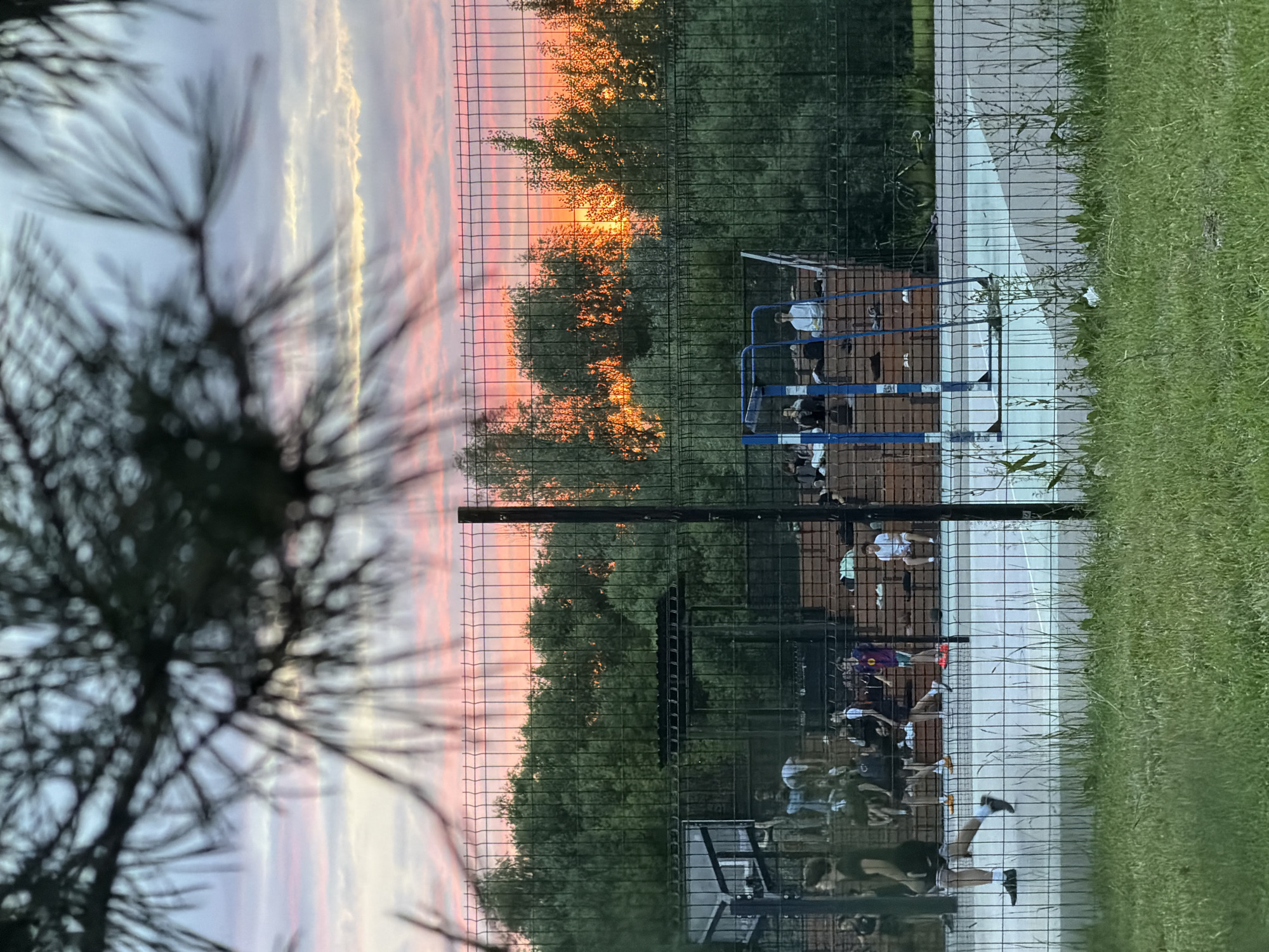 A group of people are gathered at an outdoor basketball court in Kazan, Russia, at sunset. The sky is filled with vibrant pink and orange clouds as the sun dips below the horizon, casting a warm glow on the trees. The court has a fenced-in perimeter and appears to be a popular spot for recreation. Several individuals are visible on and around the court, some appearing to be playing basketball, while others are observing or resting. The overall atmosphere seems relaxed and social, with the warm light of sunset adding a peaceful ambiance to the scene. The image is captured from a slight distance, with a pine tree in the foreground partially obscuring the view.