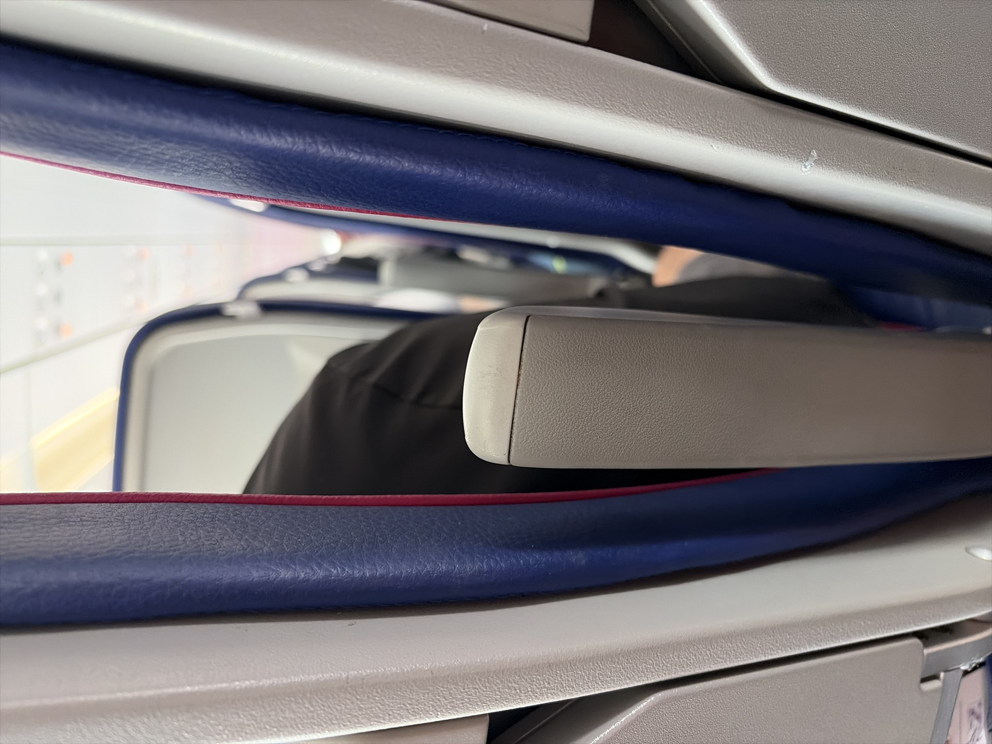 The image is a close-up, eye-level perspective of the interior of an airplane cabin, taken from a passenger's seat. The focus is on the armrests and the space between them, revealing a narrow passage.
In the foreground, the grey, textured surface of an armrest is visible on the right side, and a similar armrest is partially visible on the left. These armrests appear to be made of plastic or a similar composite material. Between them, a dark-colored object, likely the back of a seat, is seen in the middle ground, partially obscured by the armrests. This object has a textured surface.
Flanking the central armrest structure are the edges of two seats. The seat on the left has a white, textured side panel. The seat on the right has a blue, upholstered side panel with a thin pink or magenta trim. The blue upholstery has a slightly pebbled texture.
The background of the image shows a blurred perspective of the airplane cabin's aisle, with other seats visible in the distance. The lighting suggests an indoor environment, likely during flight, with overhead cabin lights visible as bright, elongated yellow shapes.
There are no specific indicators to place this image in Sofia, Bulgaria. The scene is a generic interior of a commercial aircraft.
