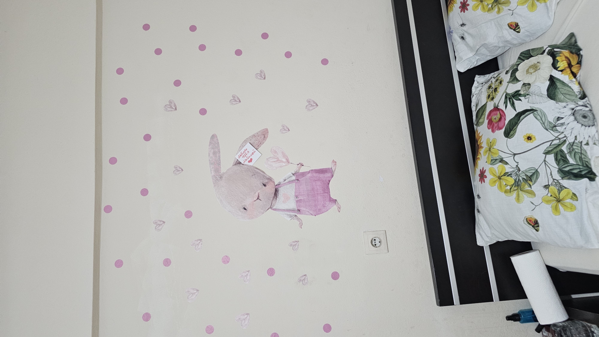 A close-up, angled shot shows a wall adorned with pink polka dots and heart decals, as well as a cartoonish illustration of a pink bunny wearing a pink vest over a white shirt. The bunny holds a heart-shaped balloon in one paw and a small tag in the other. The tag has red handwritten text that appears to be in Cyrillic script. To the right of the wall, part of a bed is visible, featuring two white pillowcases with a vibrant floral pattern and small illustrations of butterflies and insects. A black and white striped headboard or decorative panel runs vertically along the edge of the bed. An electrical outlet is visible on the wall. A roll of white paper towel and a small blue bottle are also present in the lower right corner of the frame. The setting appears to be a bedroom, likely in Maltepe, Turkey, given the location context. The lighting suggests it is daytime.
