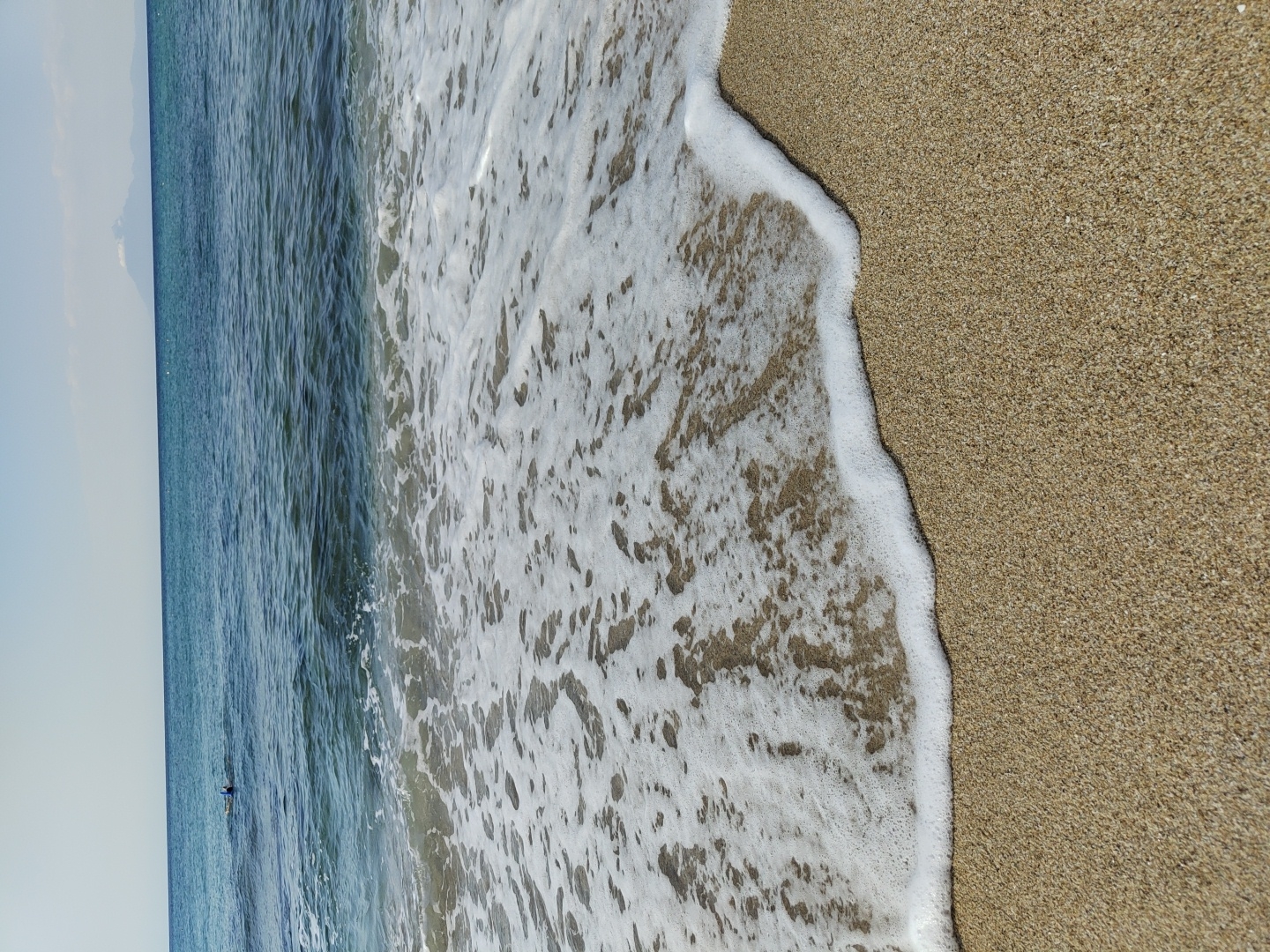 A person wearing a blue snorkel mask and fins swims in the clear turquoise waters of the Aegean Sea, approximately 50 meters from the shore of Limenaria, Greece. The person is positioned in the upper left quadrant of the frame. The sea, a gradient of deep blue to lighter turquoise, occupies the upper two-thirds of the image. Gentle waves with white foam are rolling onto a sandy beach, which fills the bottom third of the frame. In the far distance, a faint mountain range with a snow-capped peak is visible on the horizon. The sky is a pale, clear blue with a few wispy clouds. The scene evokes a sense of tranquility and a typical summer day at the beach.
