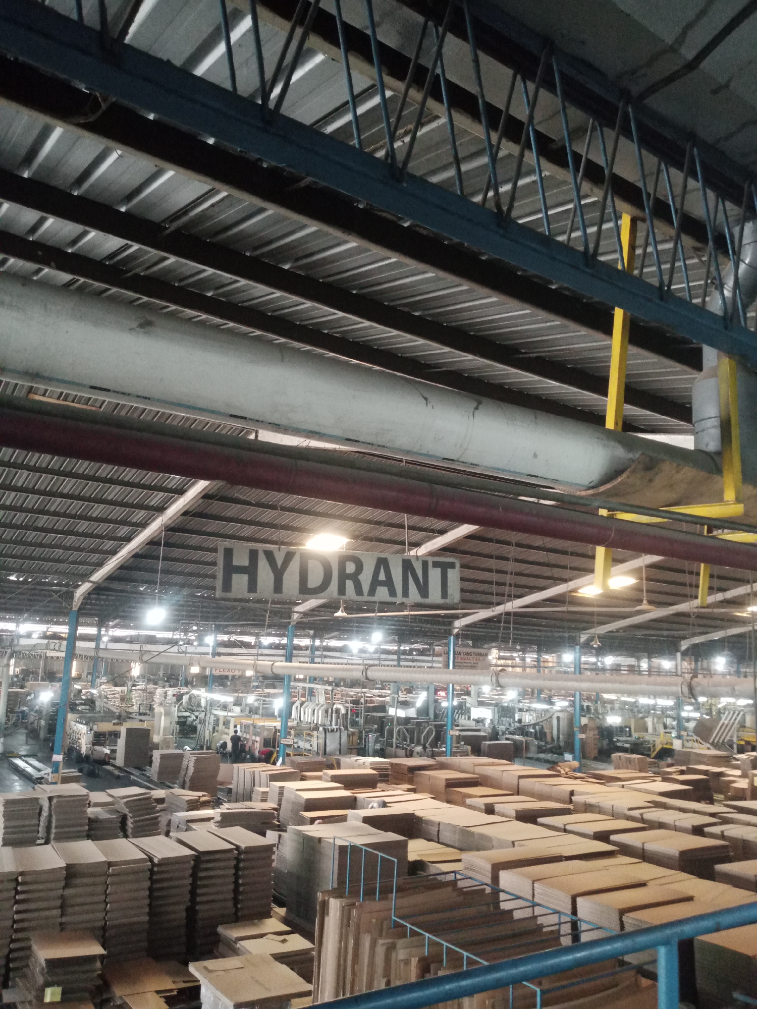 The image depicts the interior of a large, industrial facility, likely a factory or warehouse, in Balaraja, Indonesia. The ceiling is high, supported by a metal truss system, and features corrugated metal roofing. Large pipes run horizontally across the ceiling, some red, some gray. A prominent sign hanging from the ceiling reads "HYDRANT" in large, black, block letters on a white background. Below the sign, the vast space is filled with stacks of cardboard boxes of various sizes, arranged in neat rows. In the background, industrial machinery is visible, with some workers present, suggesting ongoing operations. The lighting appears to be a mix of overhead fluorescent lights and natural light filtering in from unseen sources. The overall impression is one of a busy, functional industrial setting.