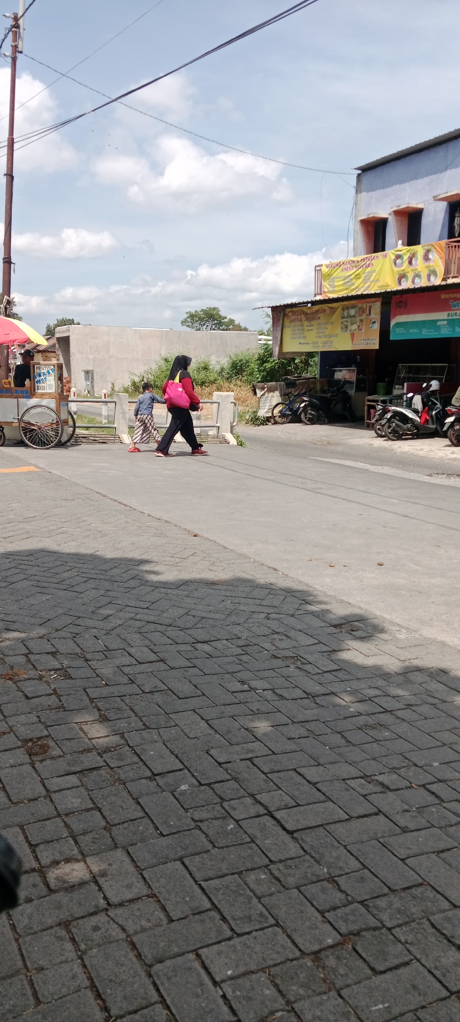 The image depicts a street scene in Maguwoharjo, Indonesia. A woman wearing a black hijab, red jacket, and black pants, carrying a pink bag, is walking across a road. Beside her, a young boy in a blue shirt and patterned pants is walking. To the left, a food cart with a yellow umbrella and a vendor is visible. In the background, there are several shops with banners, motorcycles parked, and a concrete wall. The road has a paved section in the foreground with brick-like paving stones and a grey asphalt section where the people are walking. The sky is blue with some clouds.