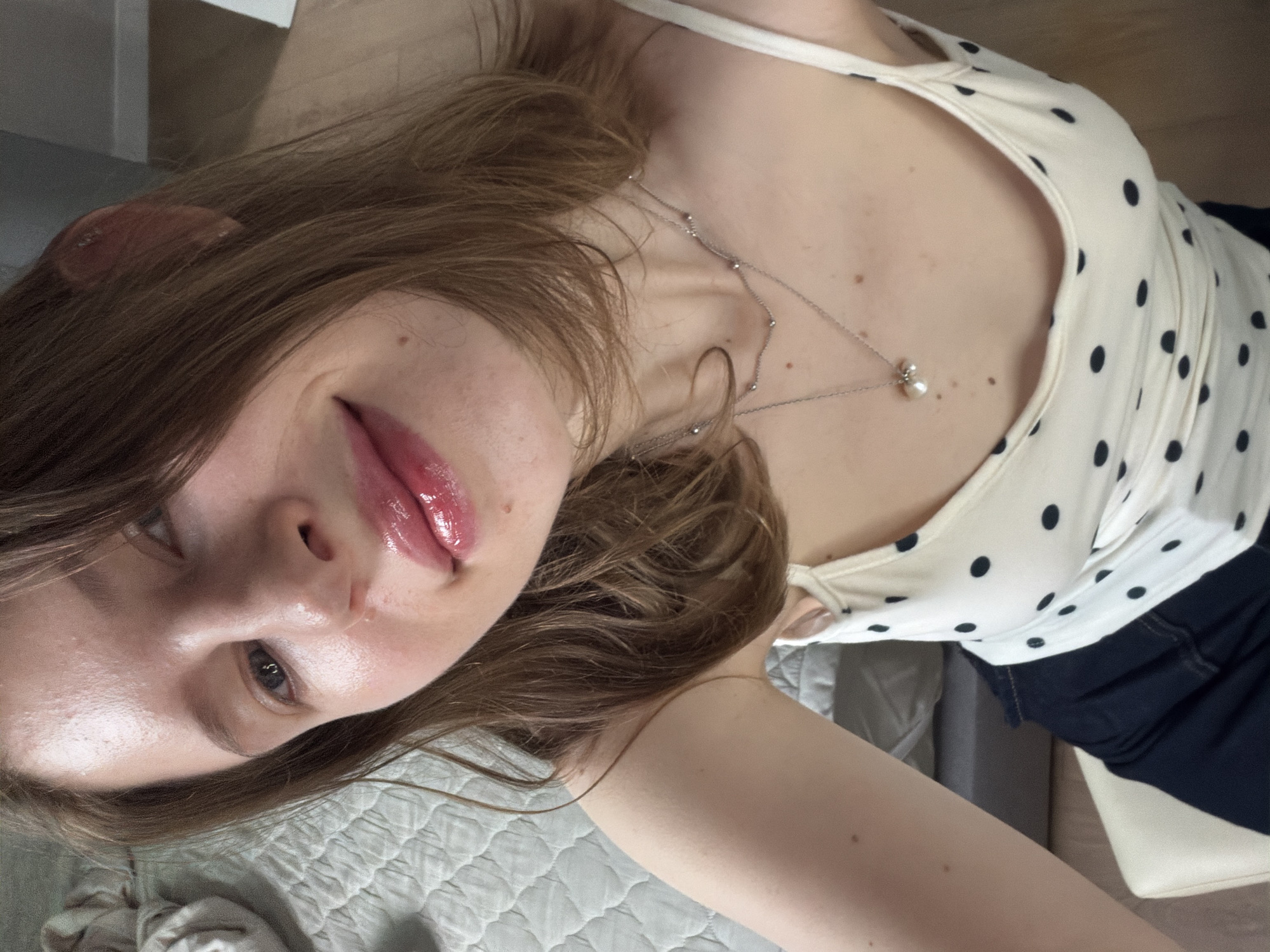 A young Caucasian female is pictured in a close-up, selfie-style shot, looking slightly off-camera. She has fair skin, medium brown hair, and is wearing a white tank top with black polka dots and a delicate silver necklace with a pearl pendant. Her lips are glossy and appear to be lightly colored. The background suggests an indoor setting, possibly a bedroom, with a textured blanket visible on a bed. The lighting is soft and diffused. There is no specific information to indicate the location is Dongducheon-si, South Korea.