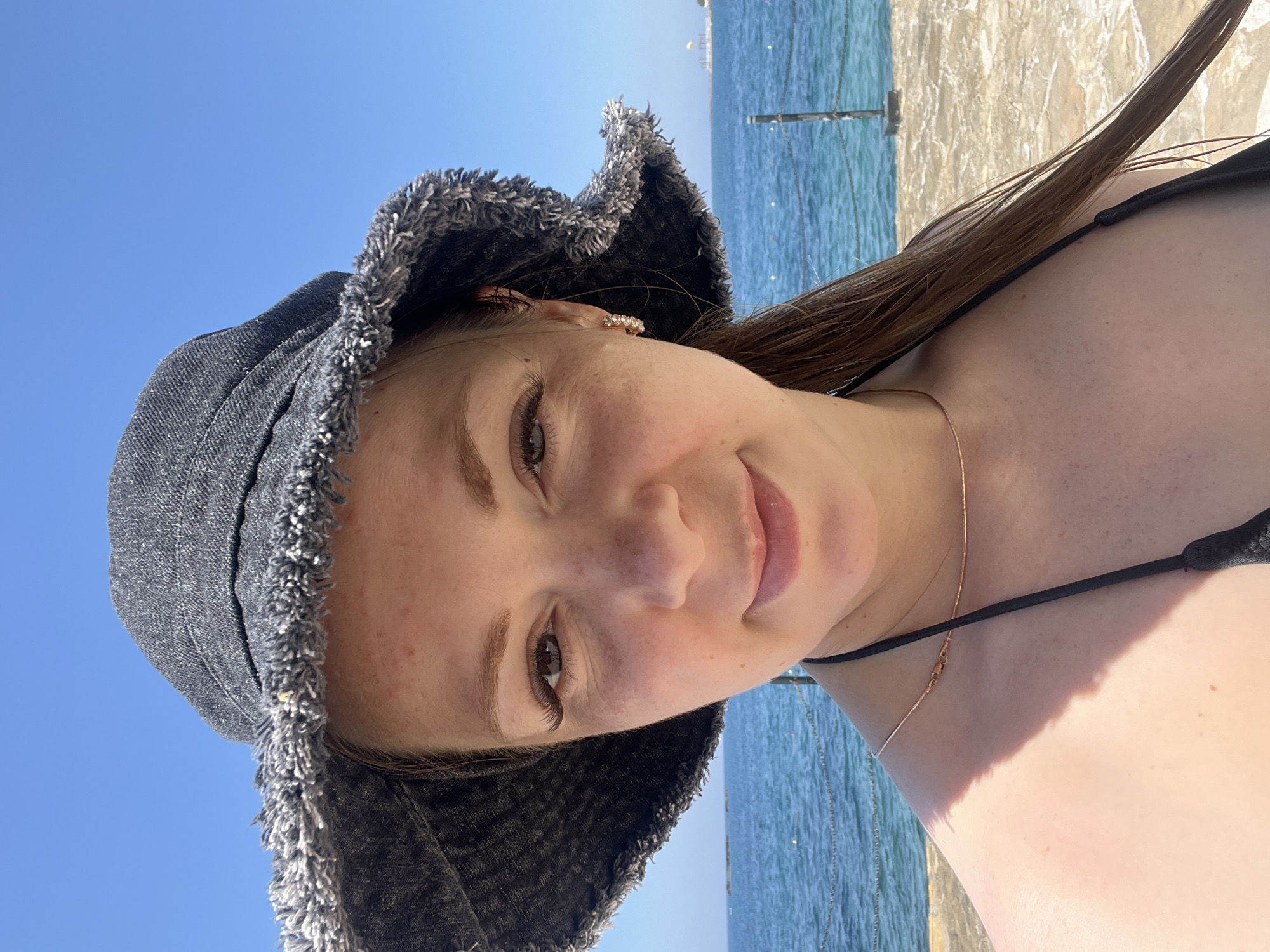 A woman with a bucket hat smiles at the camera, with the blue sea and sky behind her. She is wearing a black bikini top and a thin gold necklace. The water appears to be calm with some faint white lines suggesting small waves. A stone walkway or pier is visible in the foreground. The location is Alanya, Turkey.