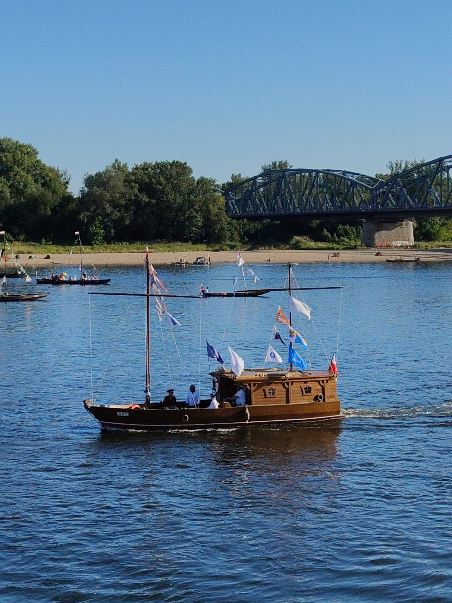 This is a medium-shot, eye-level, vertical, outdoor, daytime, natural light photograph of a historical wooden boat sailing on a body of water. The boat has a single mast with several flags flying. There are at least three people on board, two of whom are visible in the foreground, seated. The boat is moving, creating a wake behind it. The water is a deep blue with small ripples. In the background, there is a sandy shore with lush green trees and a large, dark metal bridge spanning the water. The sky is clear and blue.