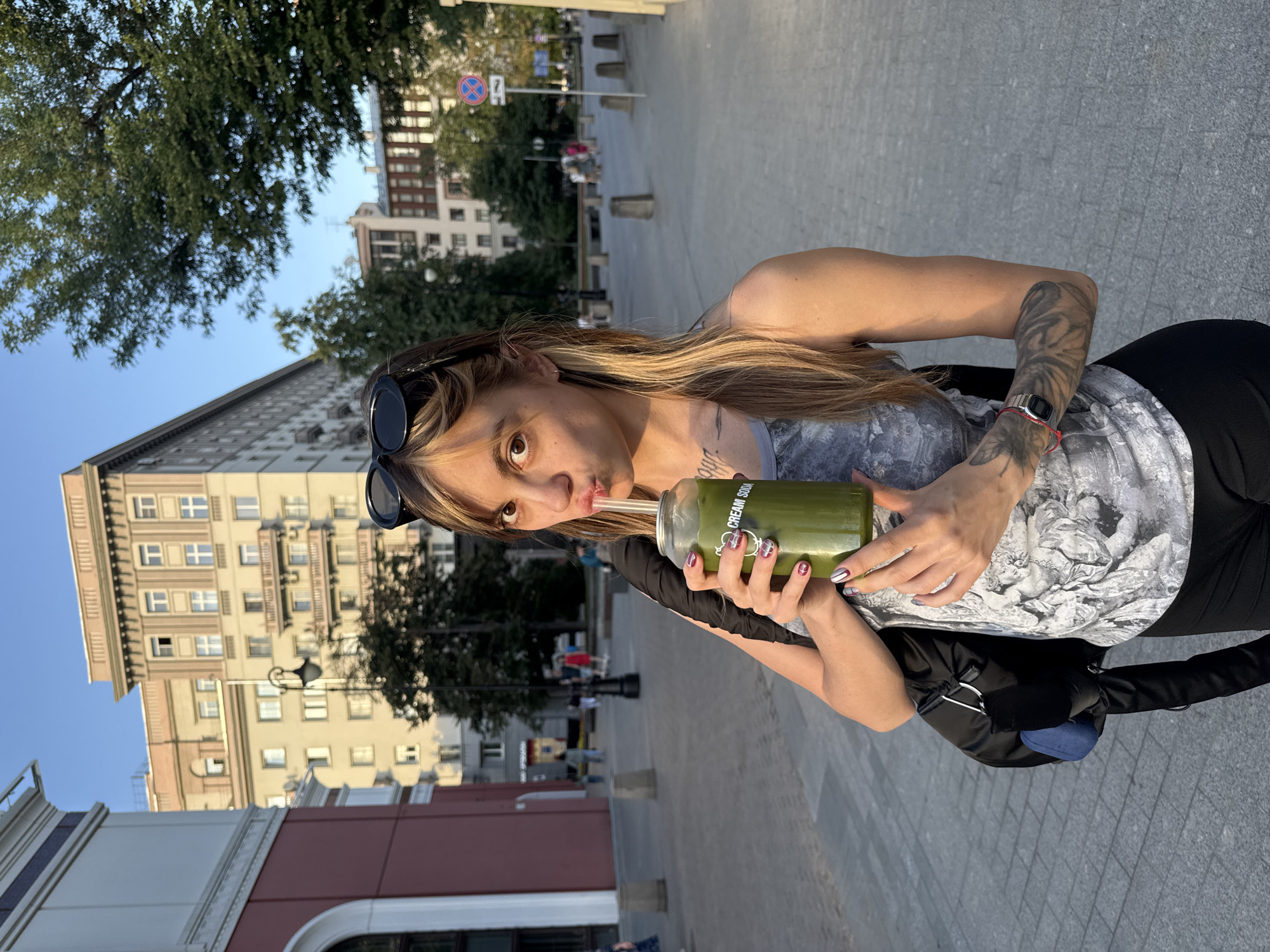 A woman with long blonde and brown hair, wearing sunglasses pushed up on her head, a gray tank top, and black pants, stands on a paved city sidewalk. She is holding a clear glass cup filled with a green liquid and a straw, which she is drinking from. The cup has a label that reads "CREAM SODA". She has tattoos on her arms and is wearing a watch on her left wrist. She is carrying a black backpack over her right shoulder. In the background, there are multi-story buildings with windows and balconies, trees, and other people walking on the sidewalk. The scene appears to be set in a European city, possibly Moscow, Russia, given the architectural style of the buildings. The lighting suggests it is daytime, with the sun casting a warm glow.
