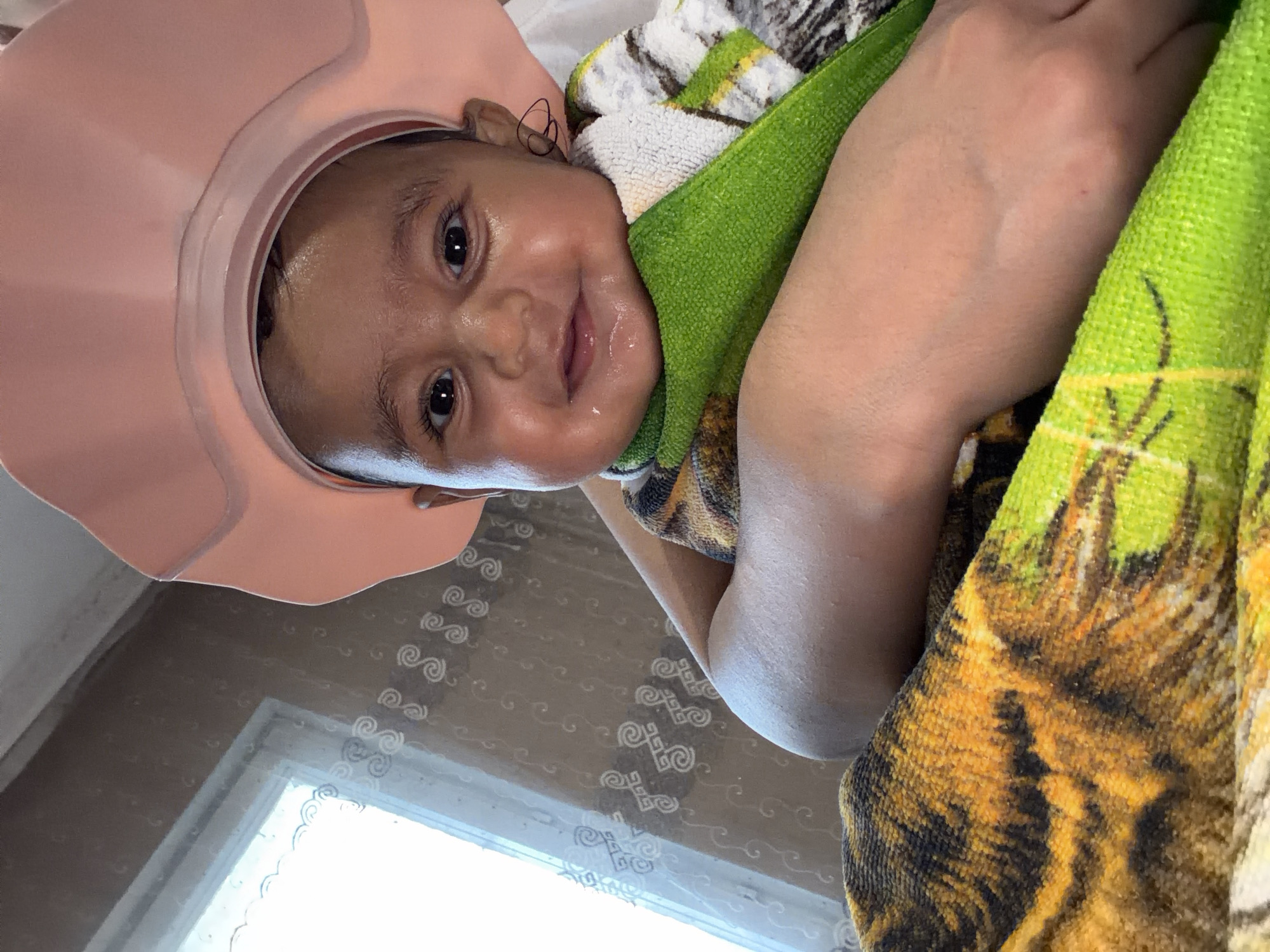 A close-up, high-angle indoor shot captures a baby wearing a pink shower cap, with its face to the camera. The baby has dark eyes and a slight smile, with moisture visible on its face. It is wrapped in a green towel and a blanket with a tiger print. In the background, a window with a white patterned curtain is visible. The scene is located in Razhevo Konare, Bulgaria.
