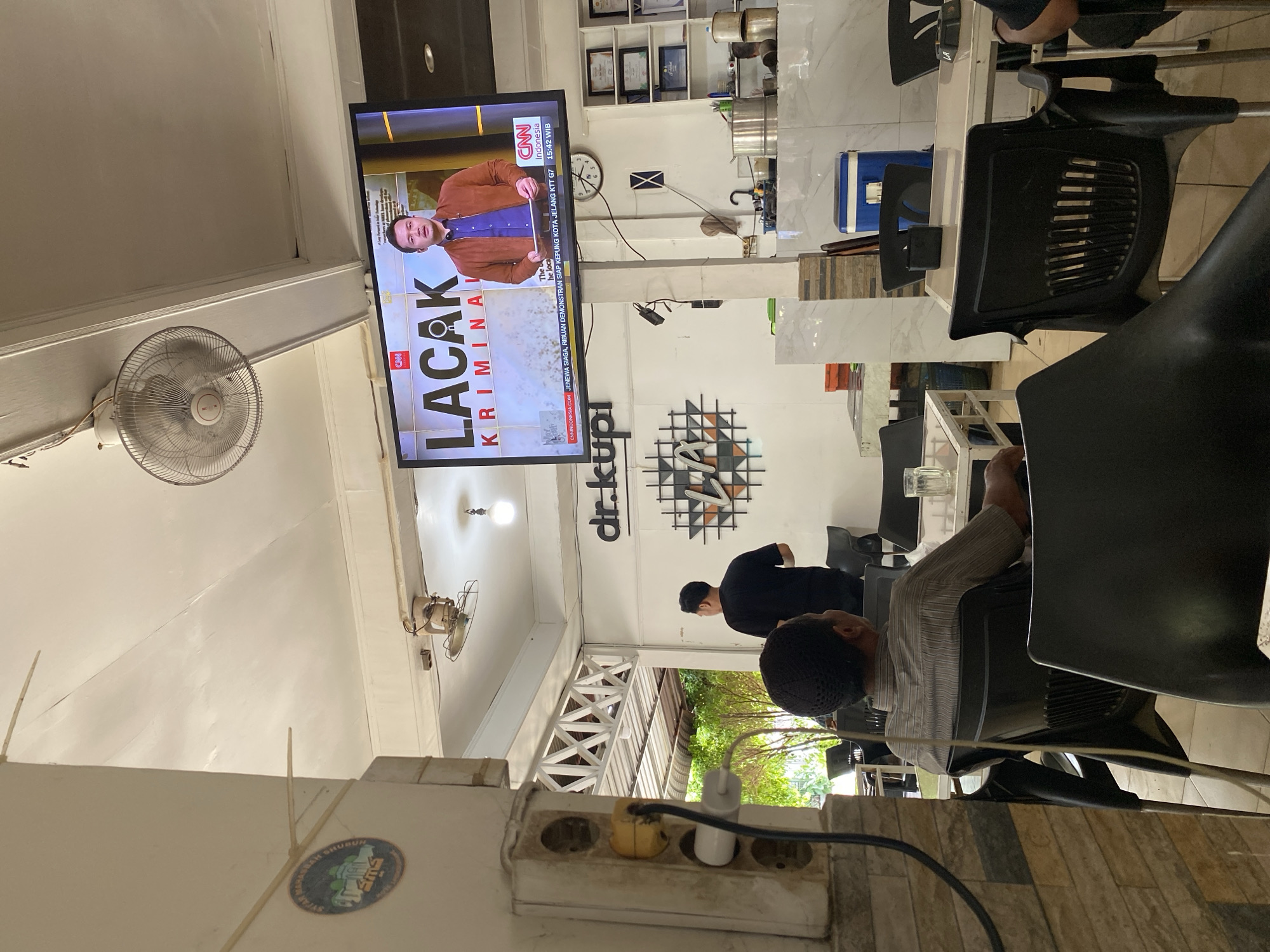 The image shows the interior of a cafe with people seated and a large television screen displaying a news program. The television shows a CNN Indonesia broadcast with the headline "LACAK KRIMINAL". A male news anchor is visible on screen. The cafe environment includes white walls, ceiling fans, tables, and chairs. In the foreground, a man wearing a striped shirt is seated, facing away from the camera. Another person in a black shirt stands further in the background. The overall scene appears to be a casual public space where people are watching television. The mention of Lhokseumawe, Indonesia, in the prompt is not visually supported by any explicit details within the image itself.